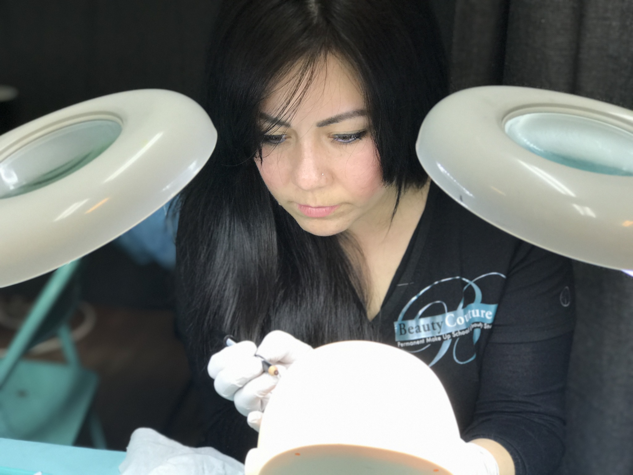 Woman with dark hair working at a table with two circular lights overhead, focused on her task.