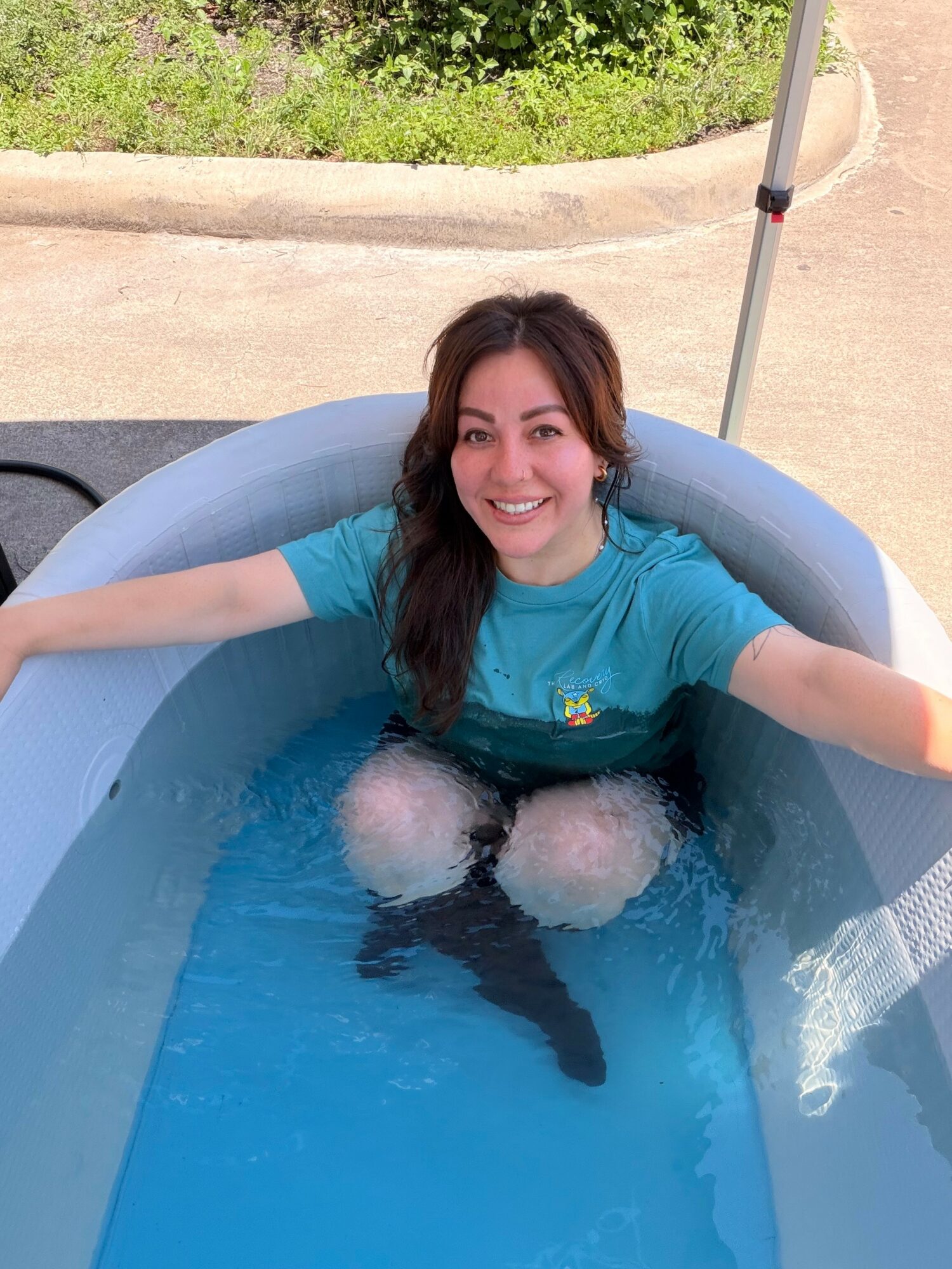 Smiling woman in a blue shirt sitting in an inflatable pool outdoors, arms outstretched, with greenery and a pole in background.