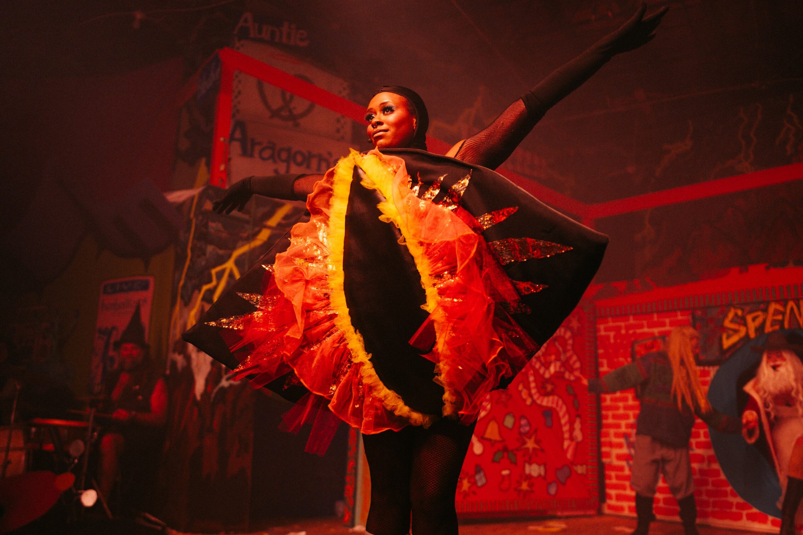 Performer in a colorful costume on stage with arms raised, surrounded by circus-themed decorations.