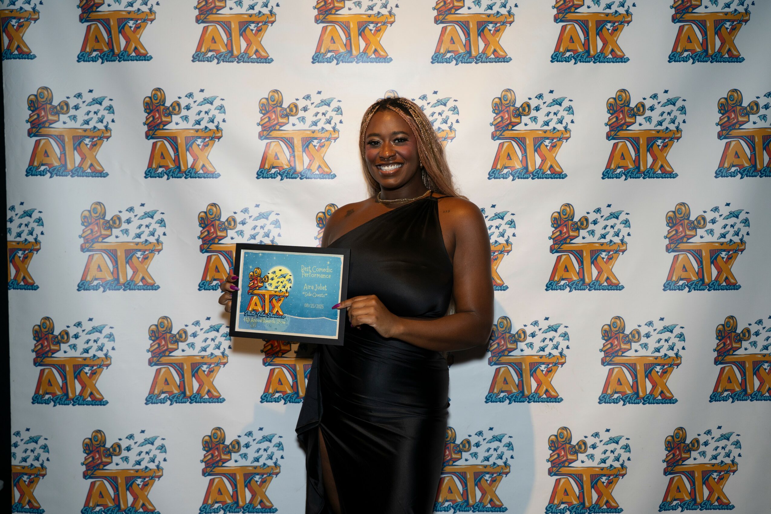 Woman with curly hair in black dress holding a plaque, standing in front of a decorated backdrop with logos.
