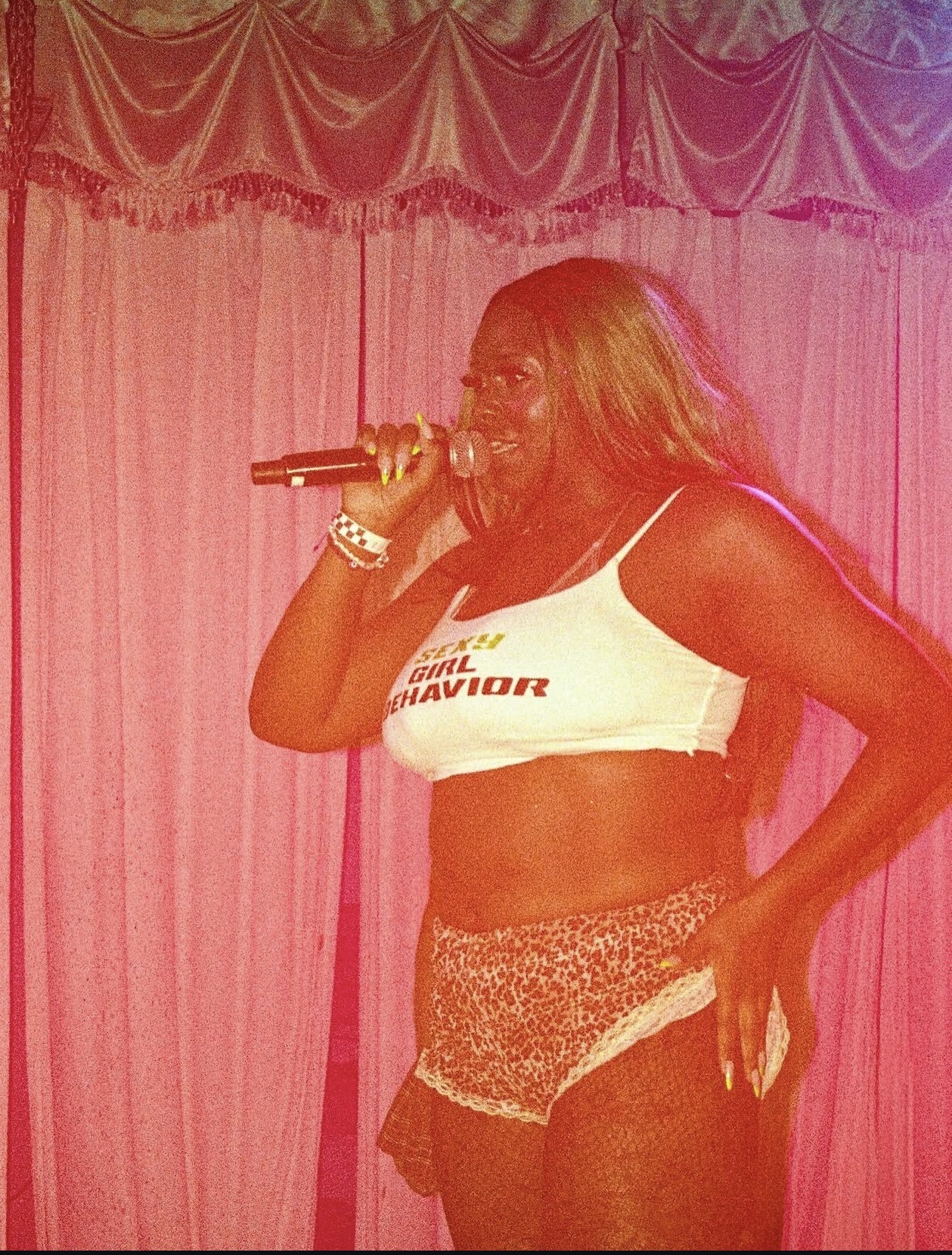 Woman singing into microphone on stage with pink curtain background, wearing crop top and shorts.