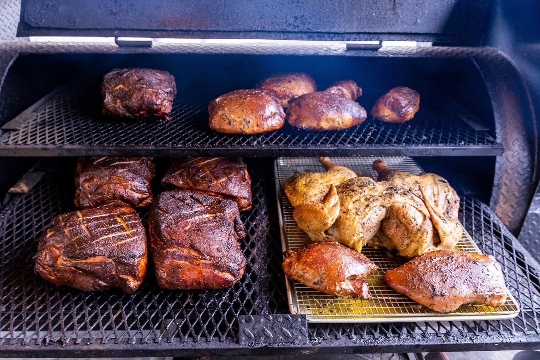 Meat and chicken pieces cooking on a grill inside an oven, with some on a tray and others on a rack.