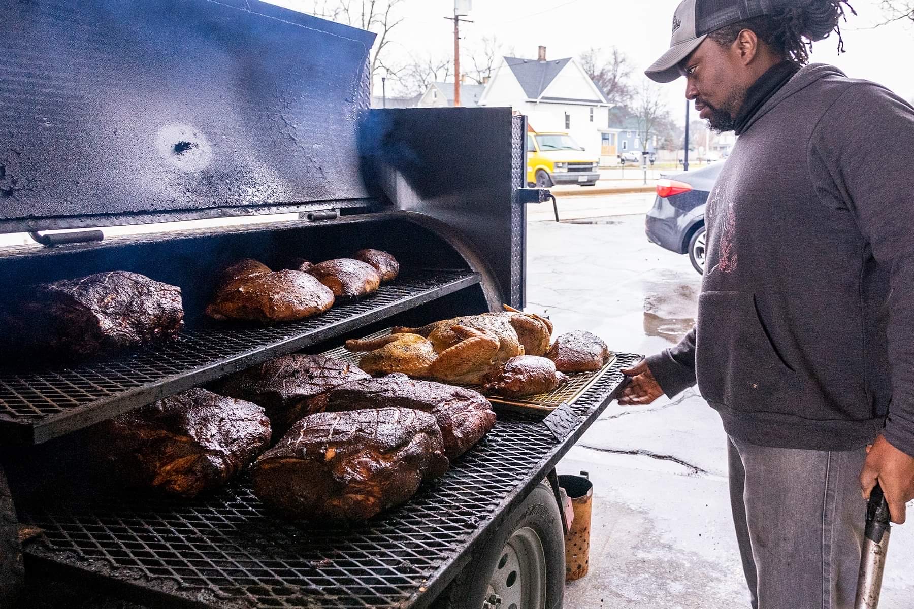 Person grilling multiple large pieces of meat outdoors on a barbecue grill, with a snowy background and houses nearby.