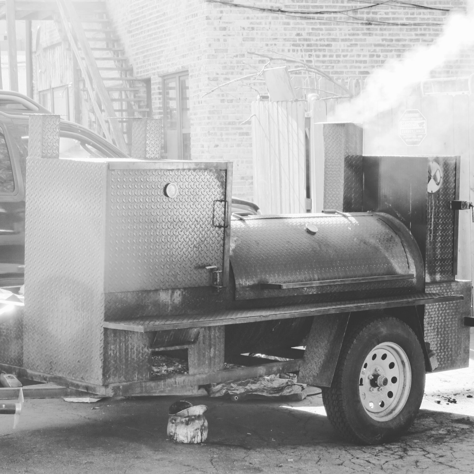 A large barbecue smoker on a trailer with a chimney emitting smoke, parked outdoors near a brick building.