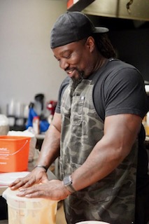 Man with a beard wearing a black cap and gray shirt, smiling while working in a kitchen.