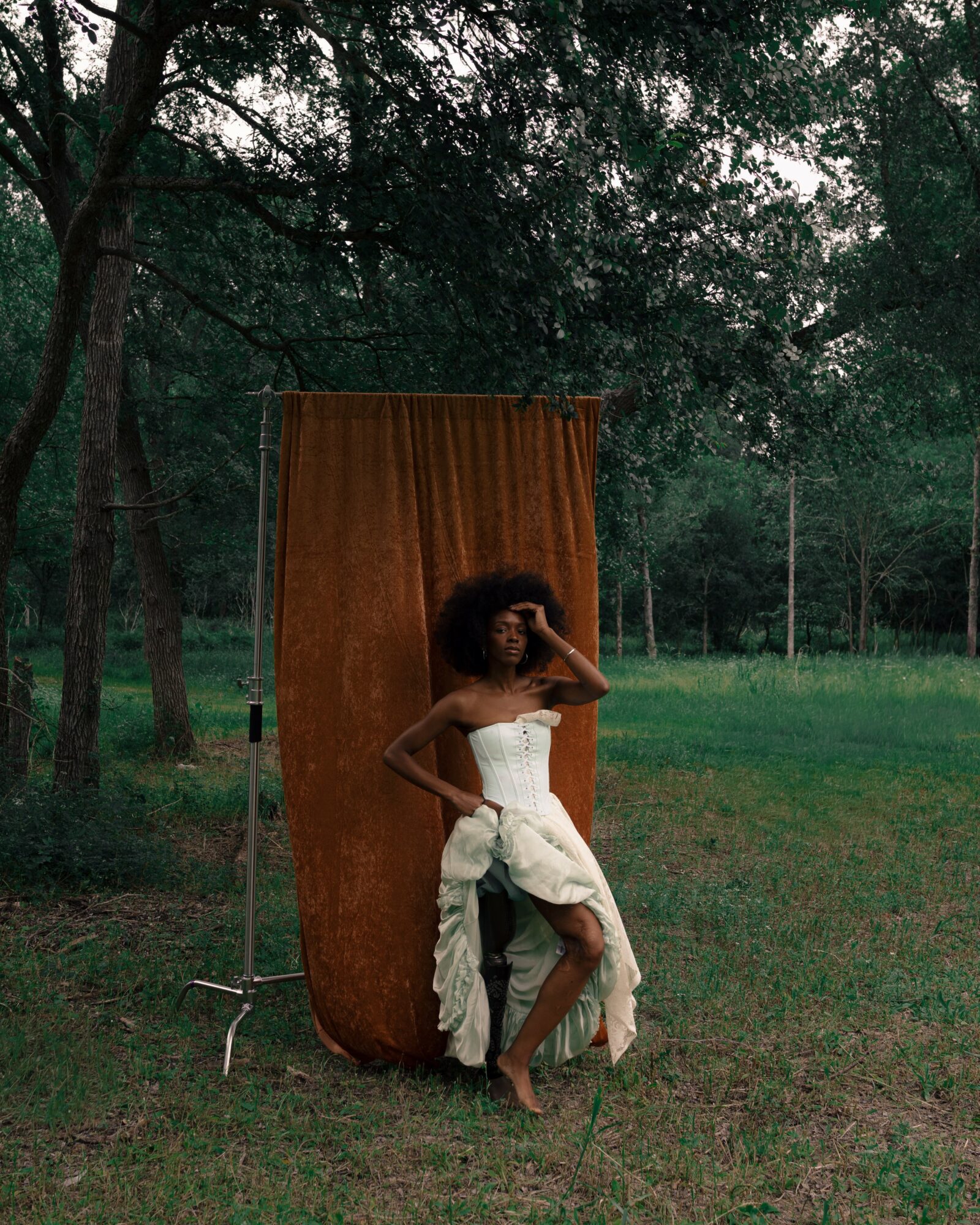 Person with curly hair in a white dress standing outdoors in front of a brown curtain, surrounded by trees and grass.