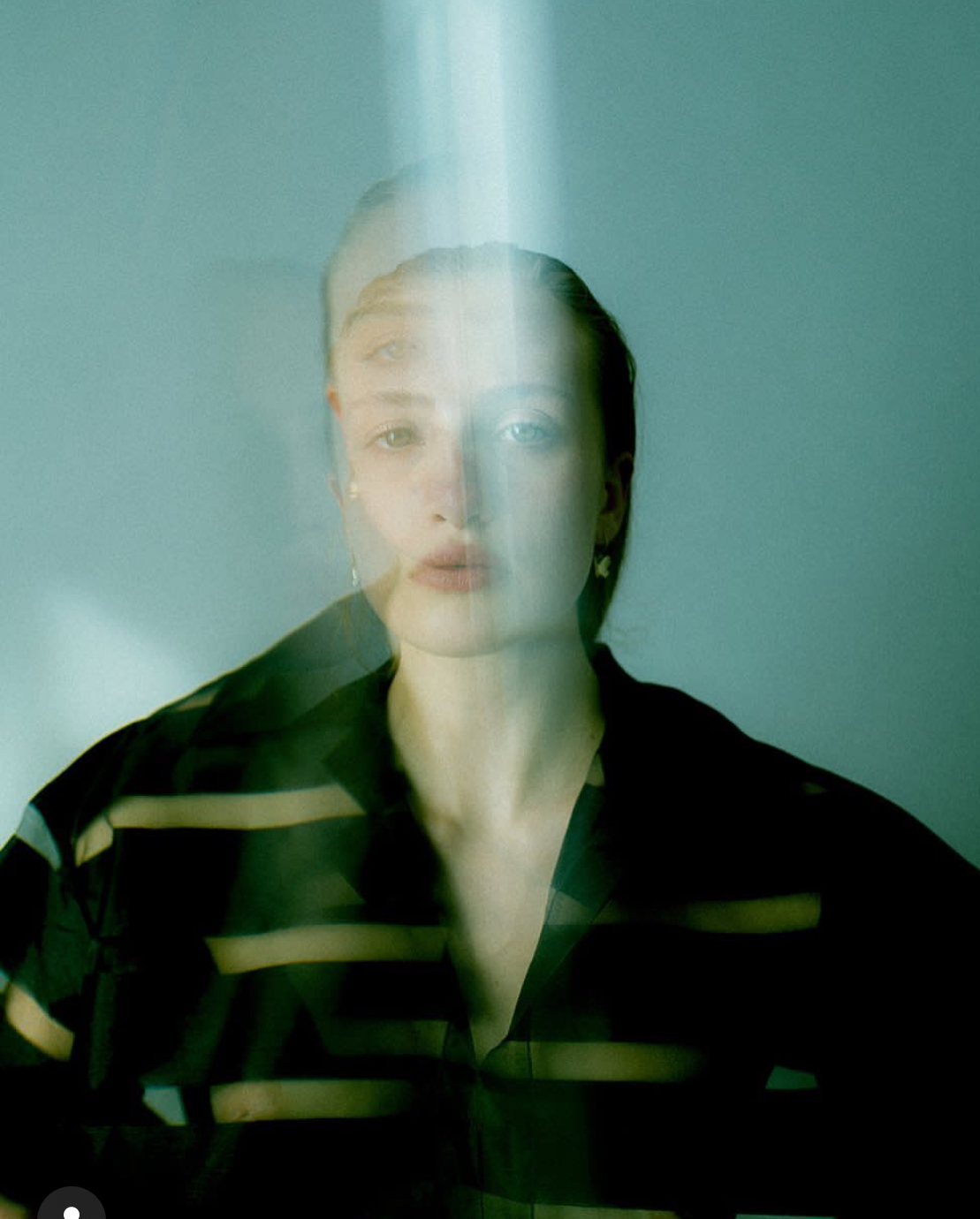 A woman with dark hair and striped shirt seen through a glass with reflections, facing forward.