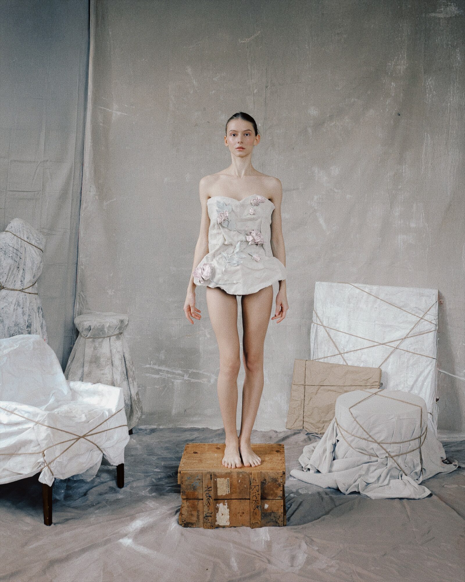 Young woman in a white dress stands on a wooden platform in a minimalist room with furniture and fabric-covered objects.