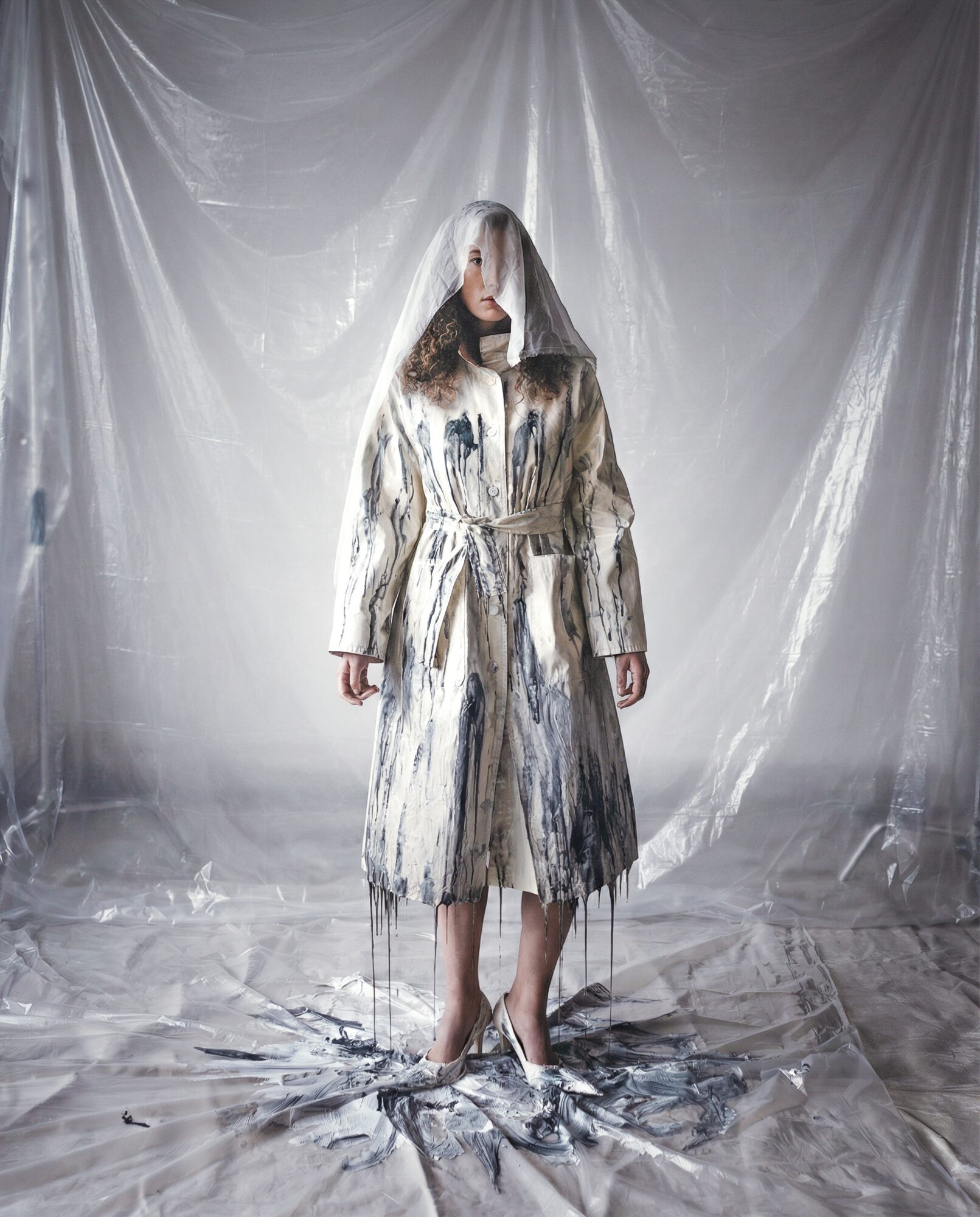 Person standing in front of a shiny, draped backdrop wearing a long, patterned coat with a hood covering their face.