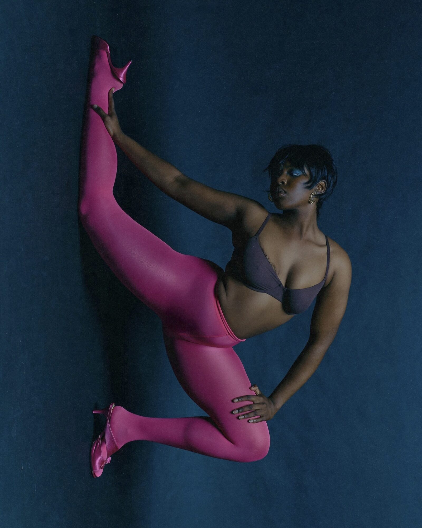 Person in black sports bra and pink leggings performs a high leg stretch against a dark background.
