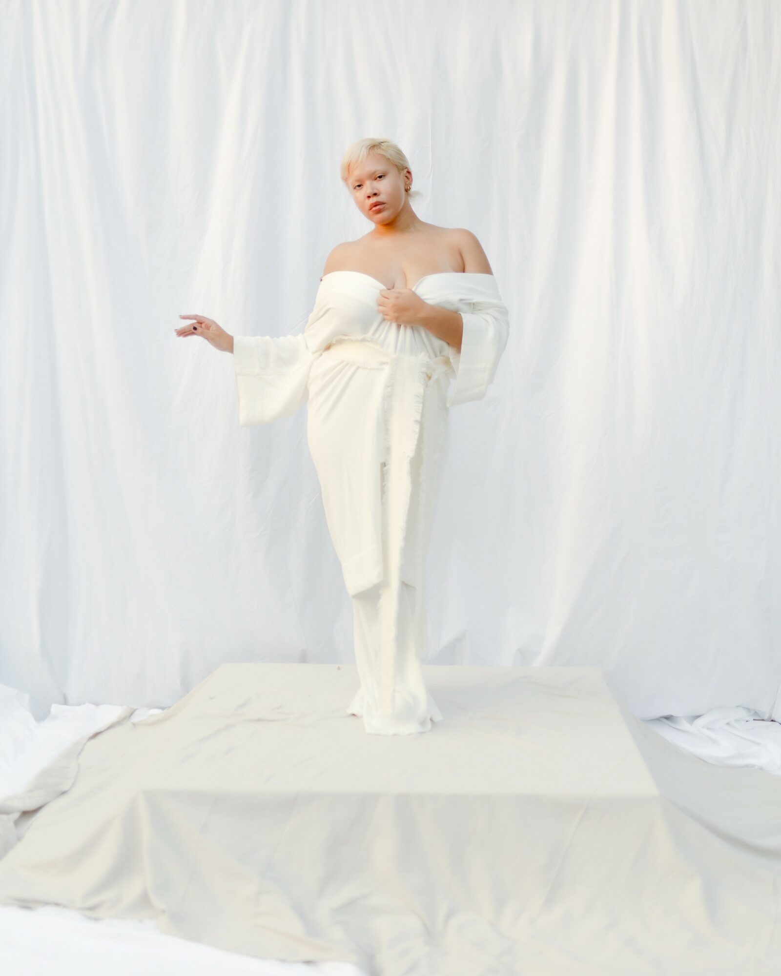 Young woman with blonde hair in a white off-shoulder dress standing on a platform against a white backdrop.
