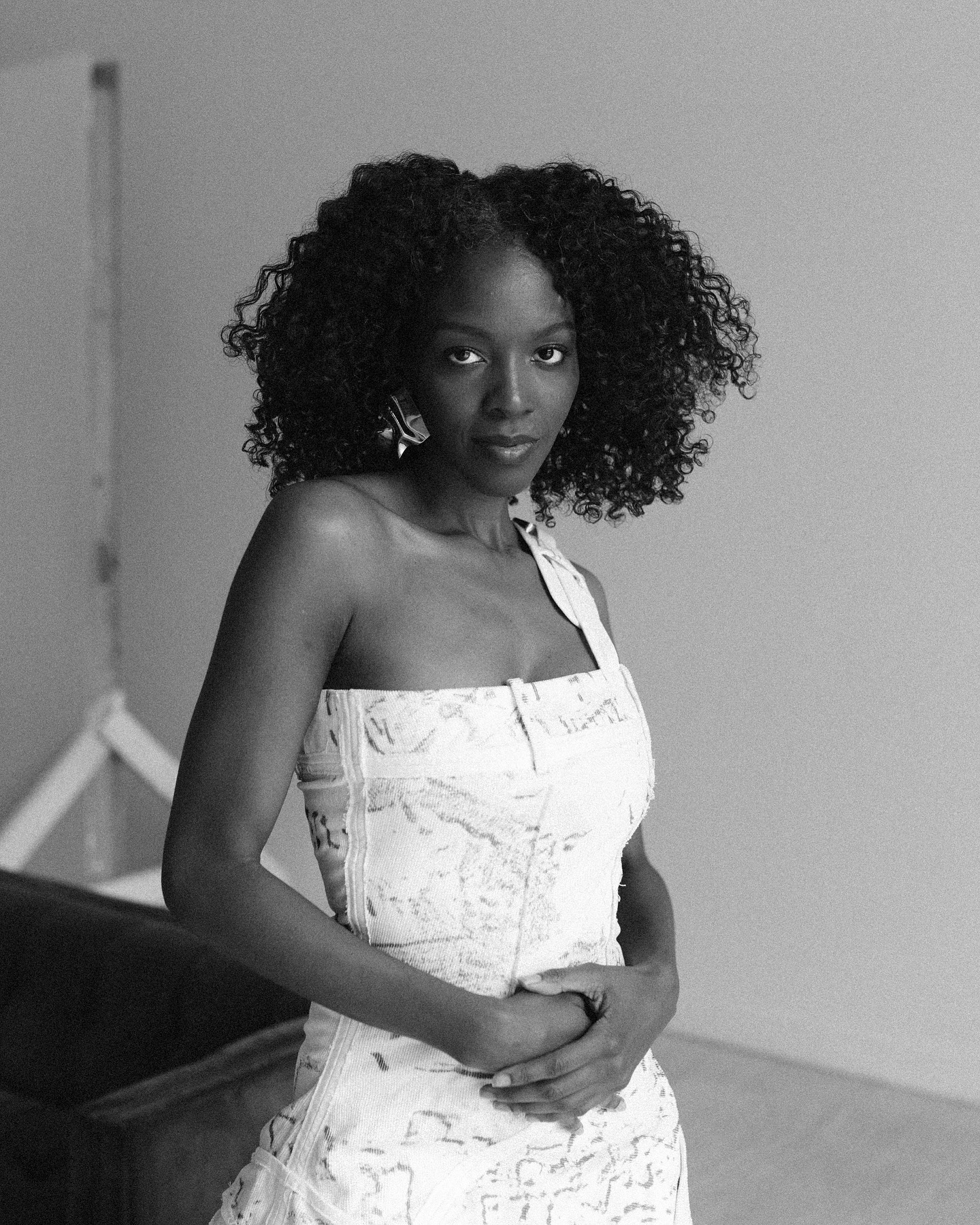 Woman with curly hair in a strapless dress standing indoors, looking at the camera, hands clasped in front.