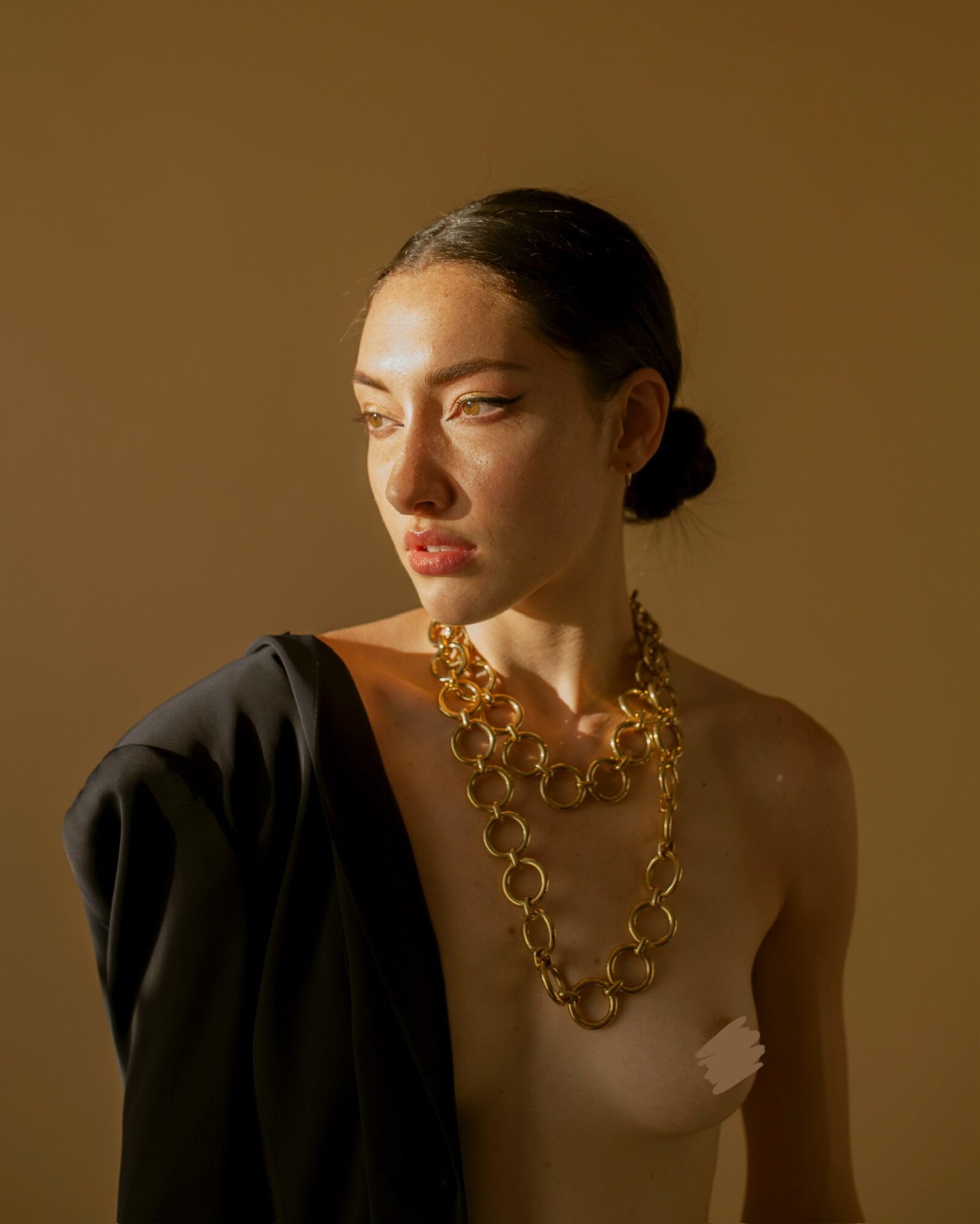 Woman with dark hair in a bun, wearing a large gold necklace, draped black fabric on her shoulder, looking to the side.