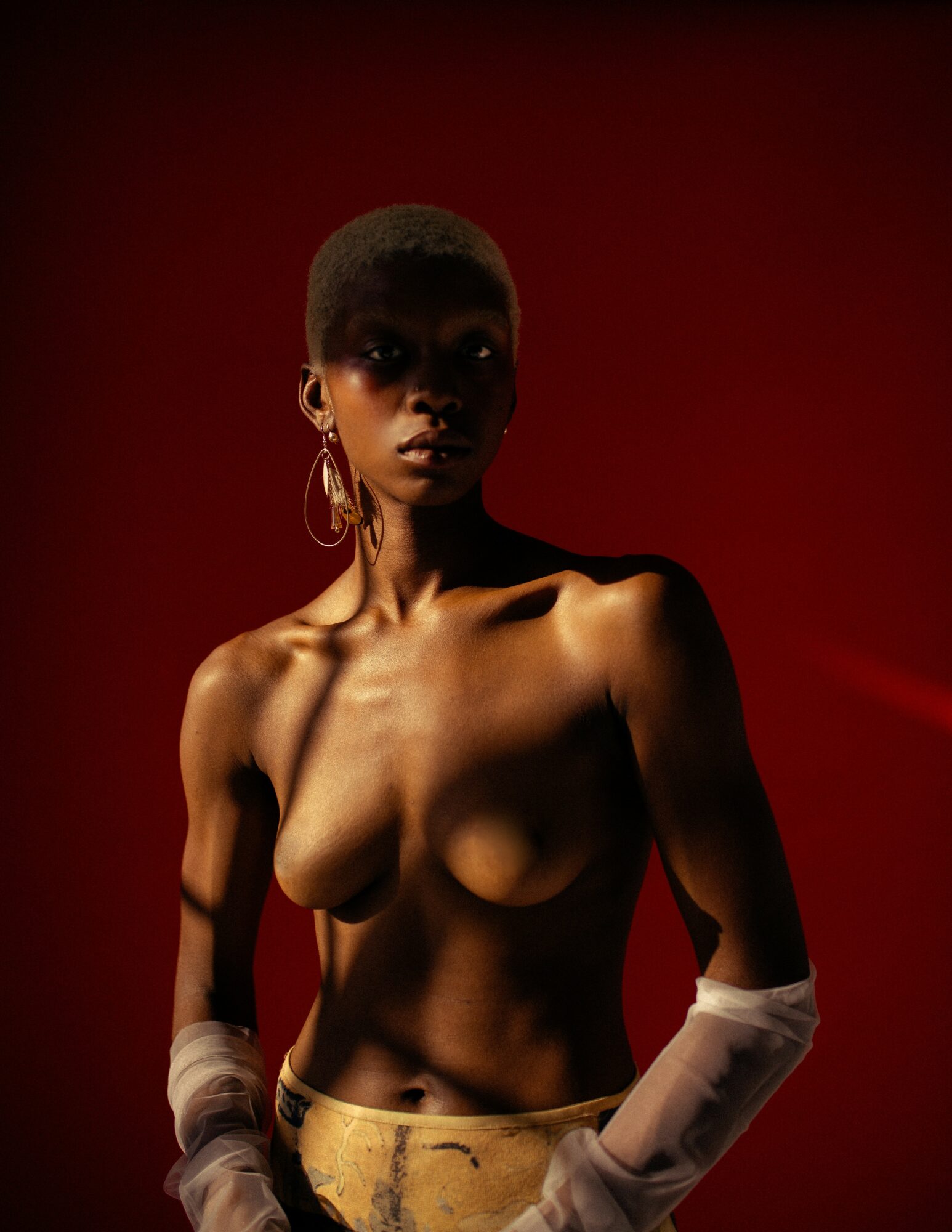 A topless woman with short hair and large earrings stands against a dark red background, with arms partially covered by sheer fabric.