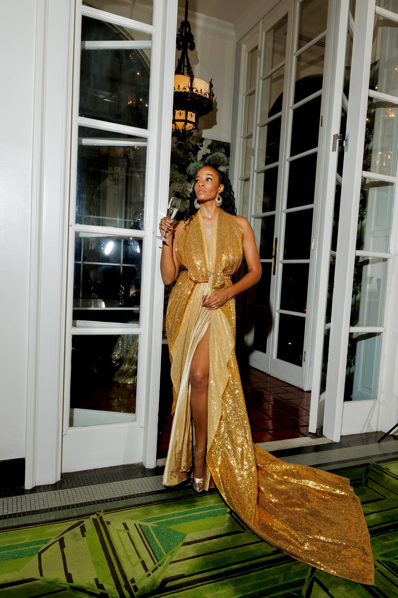Woman in a gold gown standing in doorway, holding a glass, with large windows and a chandelier inside.
