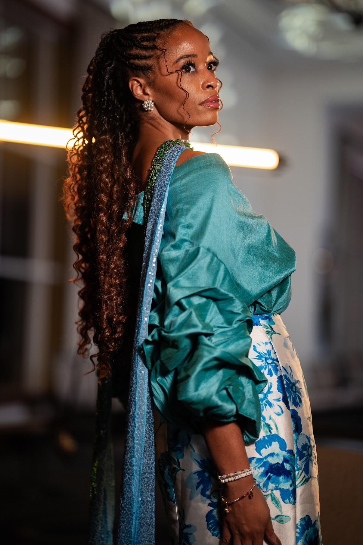 Woman with long curly hair wearing a teal jacket and floral skirt, looking to the side indoors.