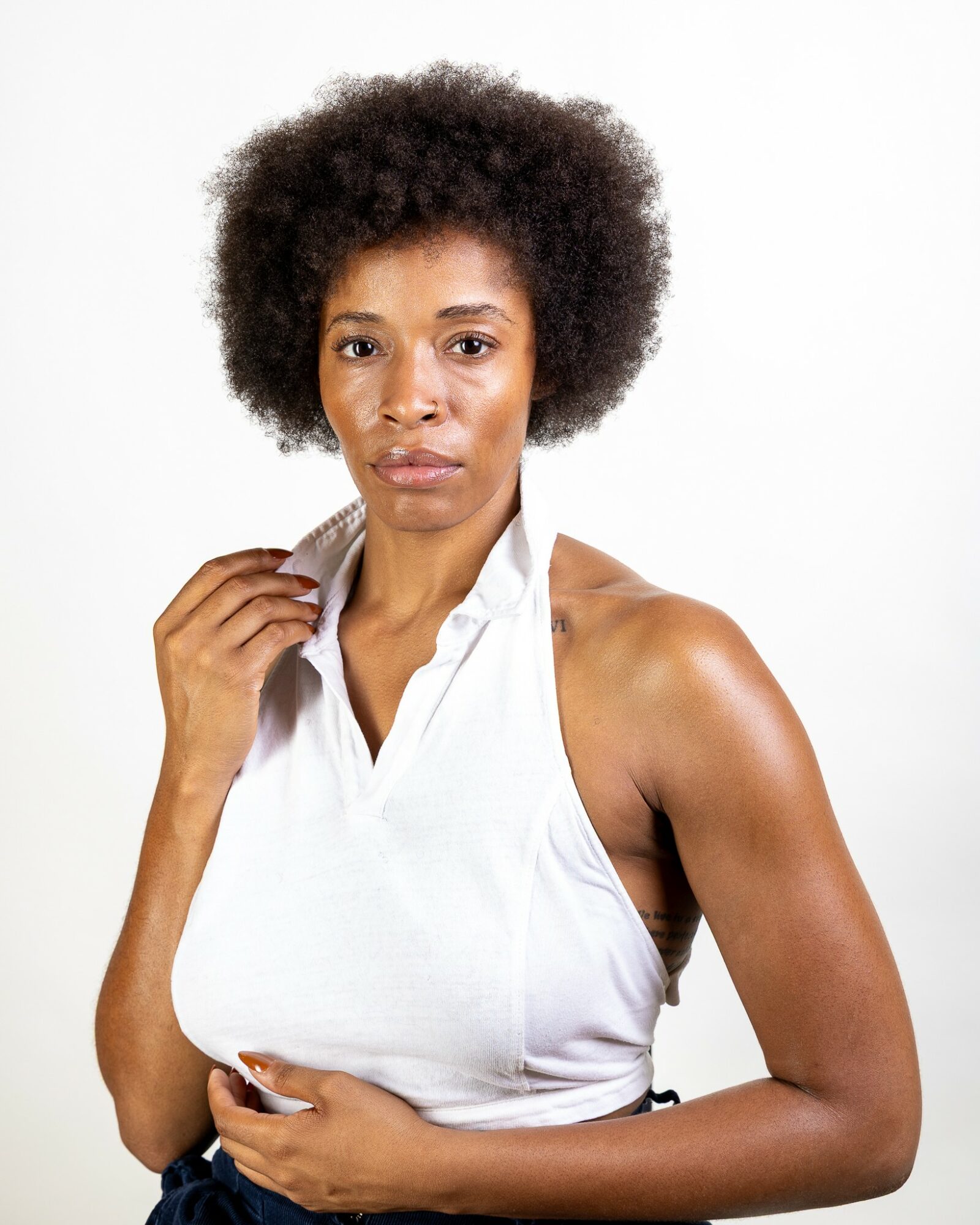 Woman with natural curly hair wearing a white sleeveless top, looking at the camera with a neutral expression.