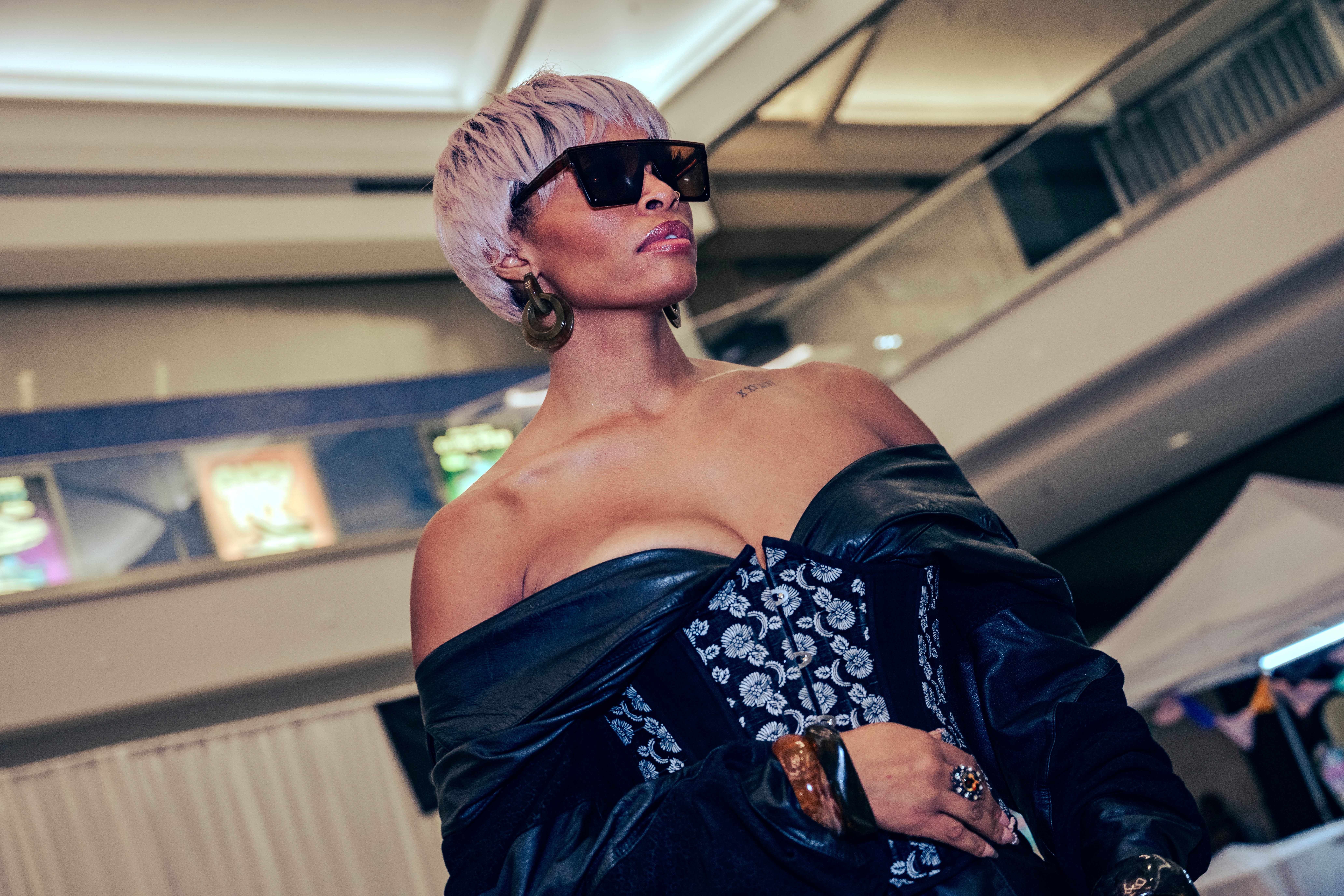 Woman with short blonde hair wearing large sunglasses and an off-shoulder black and blue dress in an indoor setting.