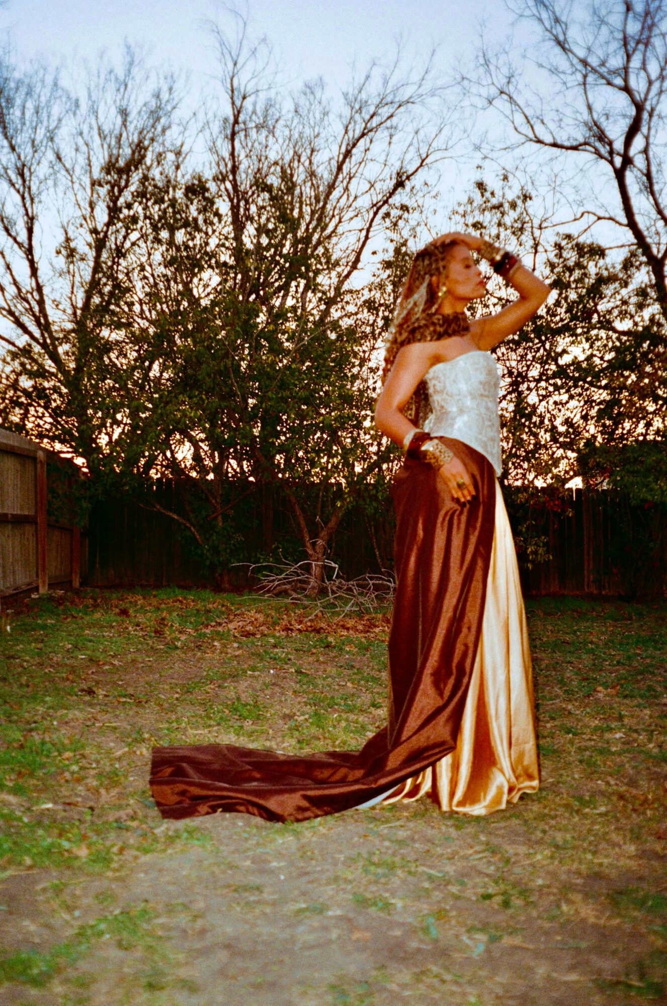 Woman in a strapless gown standing outdoors with trees and a fence, sunset sky in background.