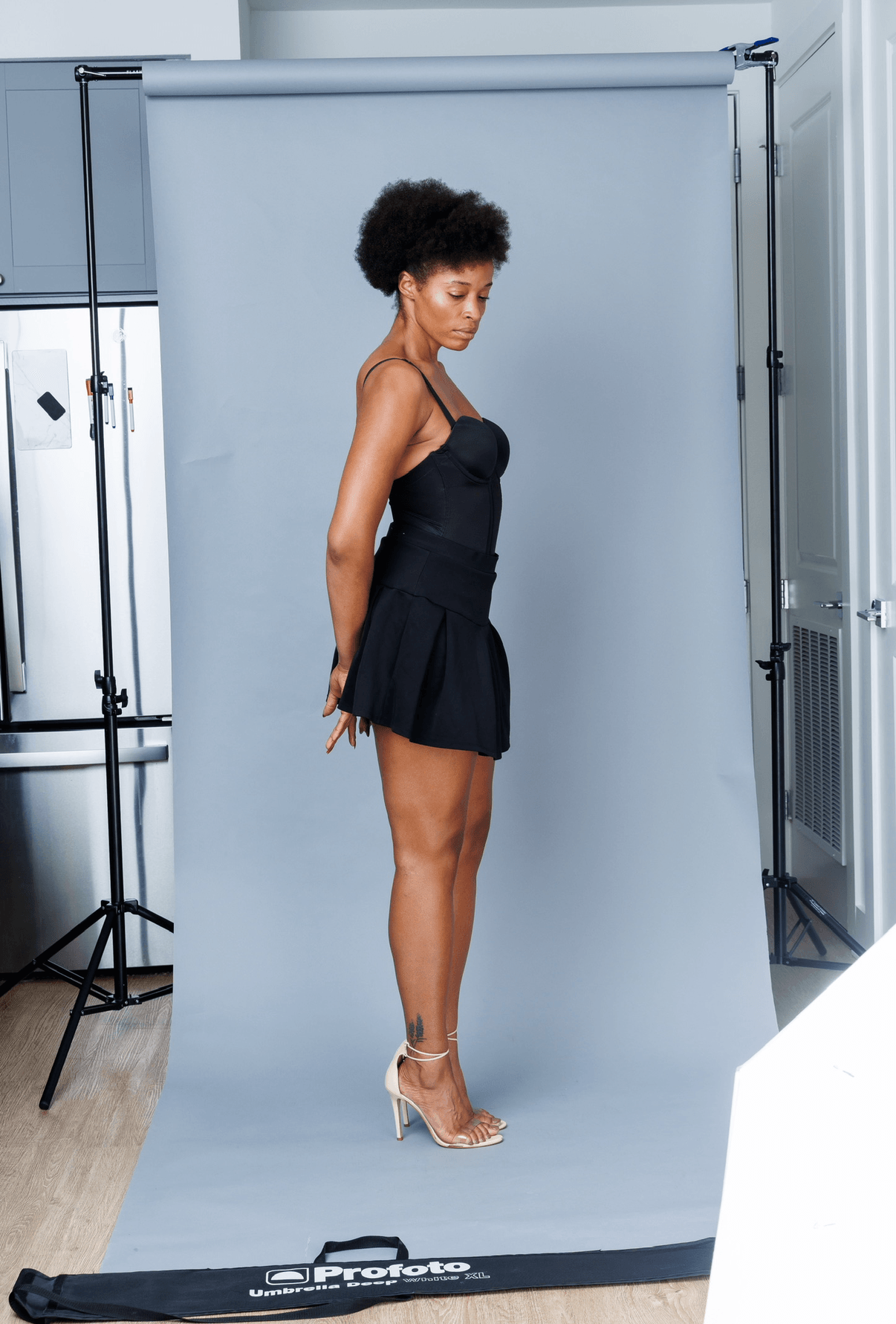 Woman in black dress and heels standing in front of a light blue backdrop, with studio equipment visible. Word count: 20