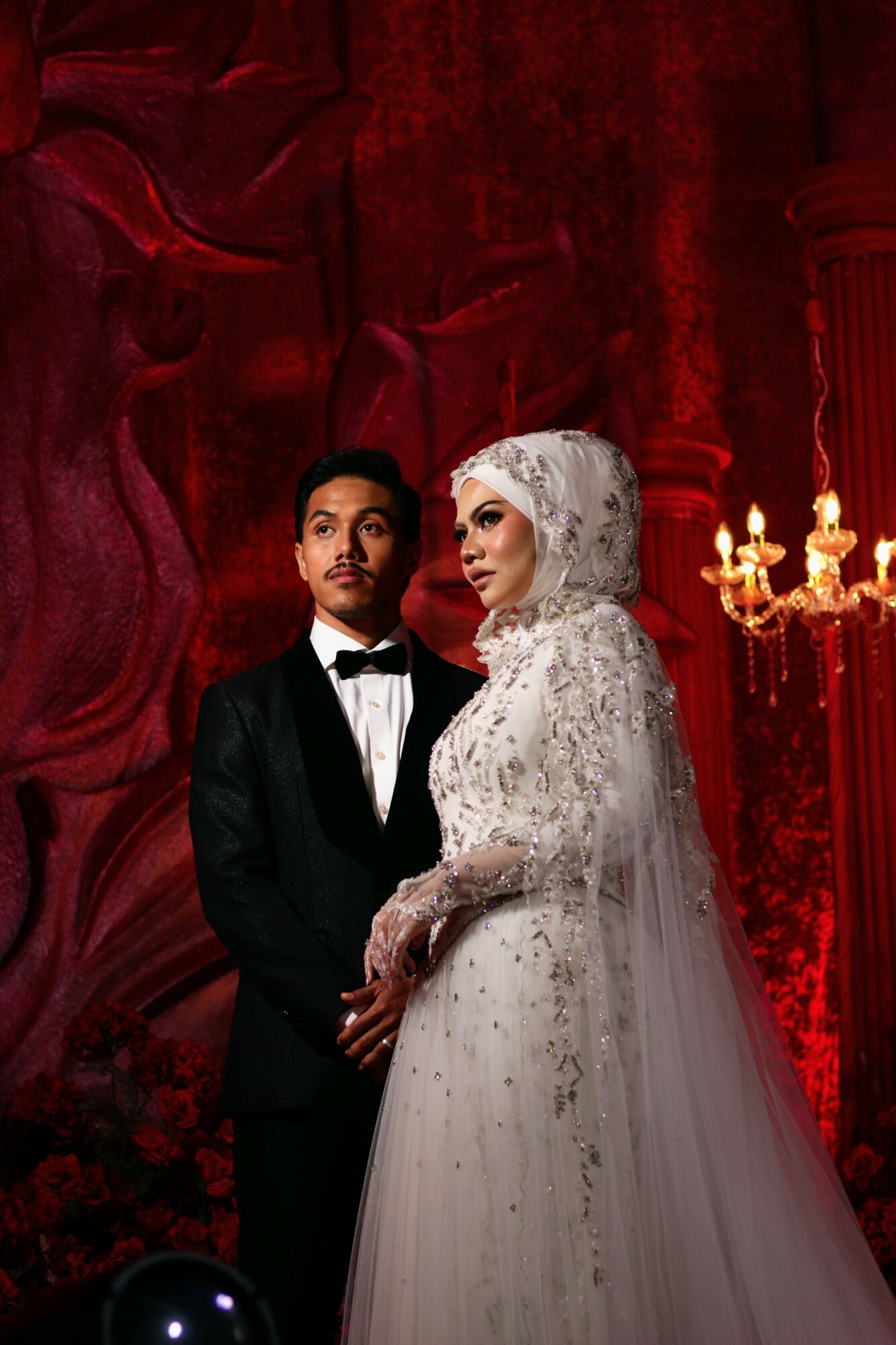 A bride and groom stand together in front of a red background with ornate decorations, the bride wearing a white lace gown and headscarf, the groom in a black tuxedo.