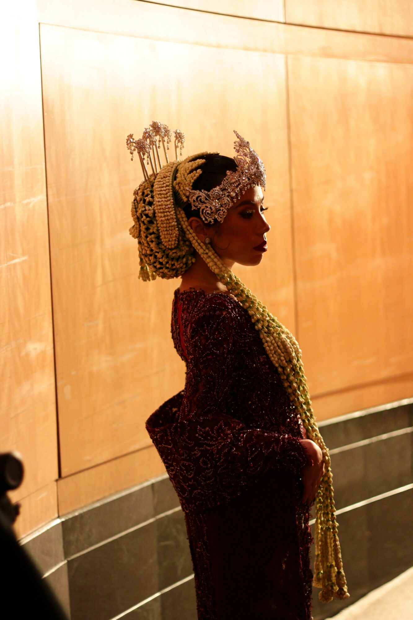 Person wearing a headscarf and a long dress, standing indoors with wooden paneling background.