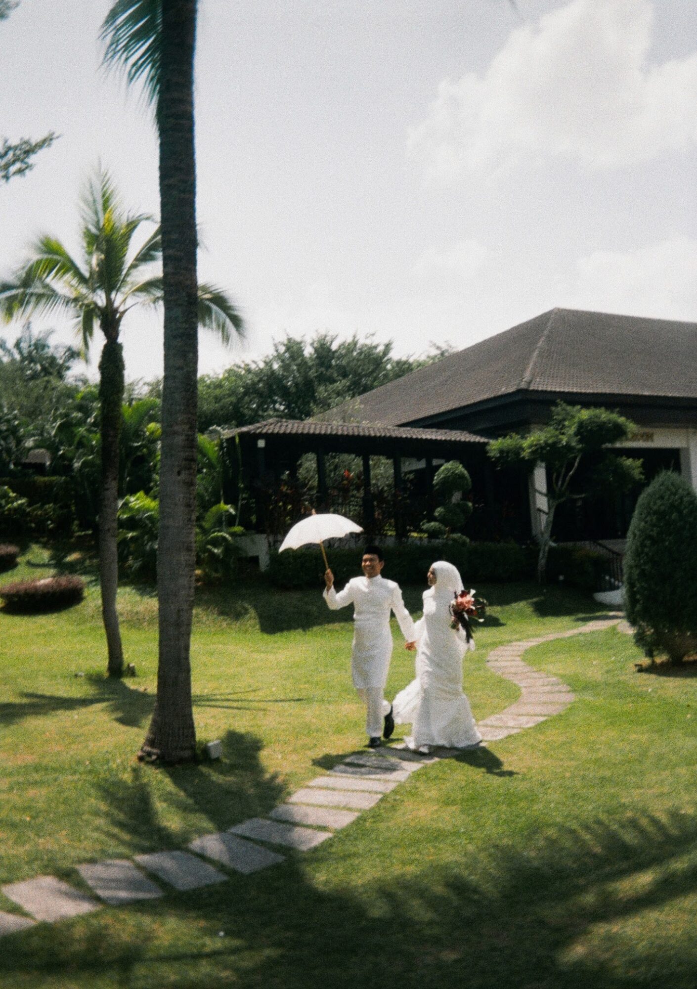 Two people in white clothing walk on a garden path, one holding an umbrella, with a house and trees in the background.