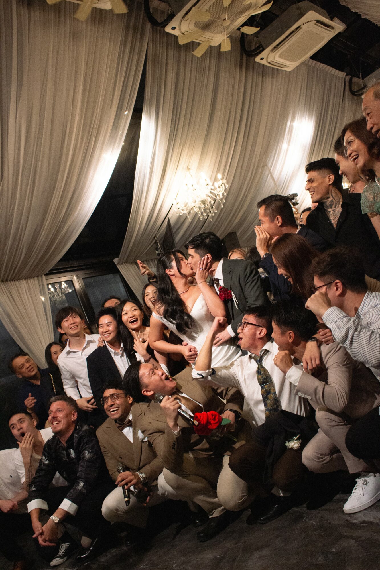 Group of people dressed in formal attire, smiling and posing together indoors with curtains and lighting in background.