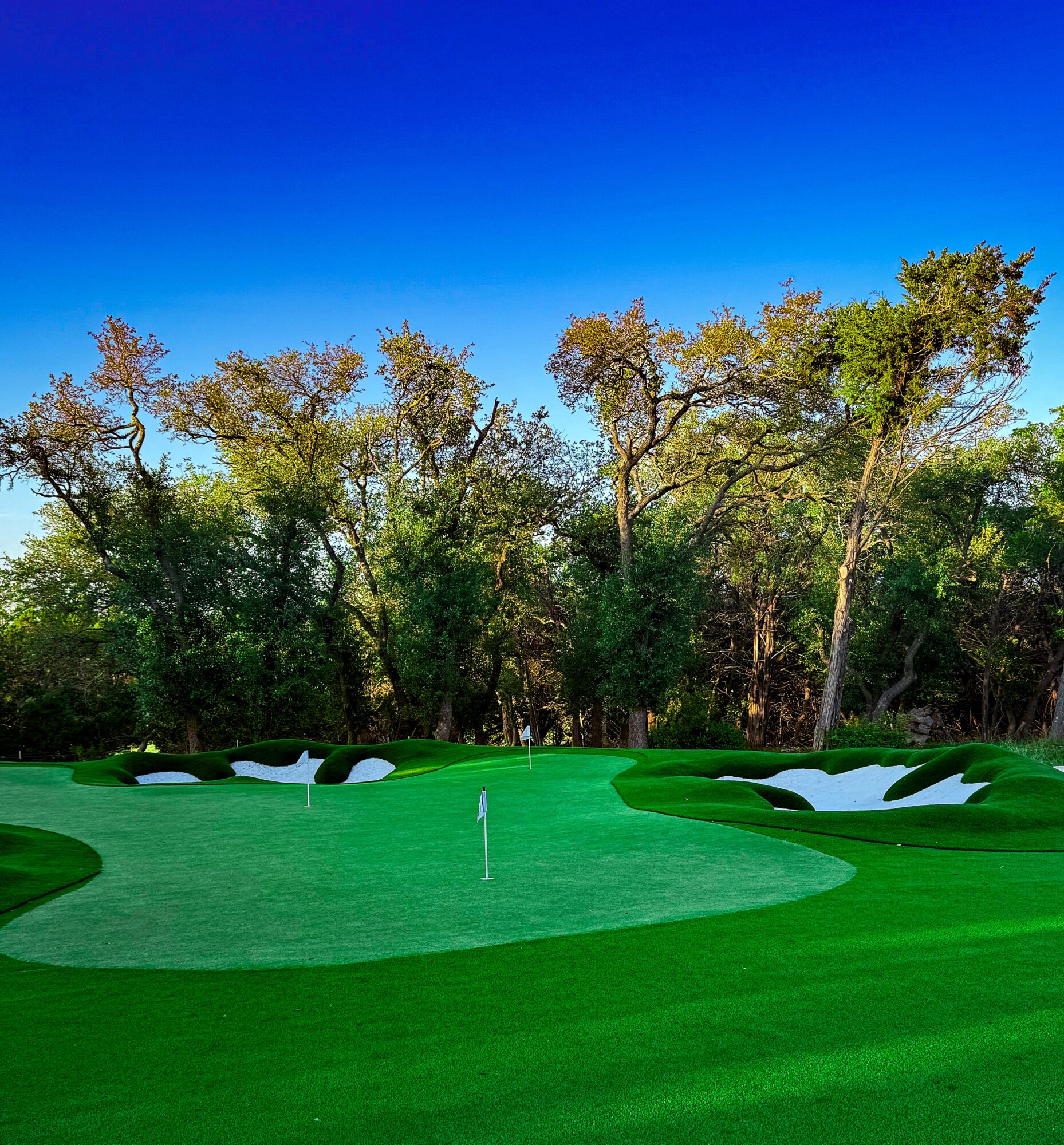 A golf green bordered by white sand bunkers and trees beneath a clear blue sky.