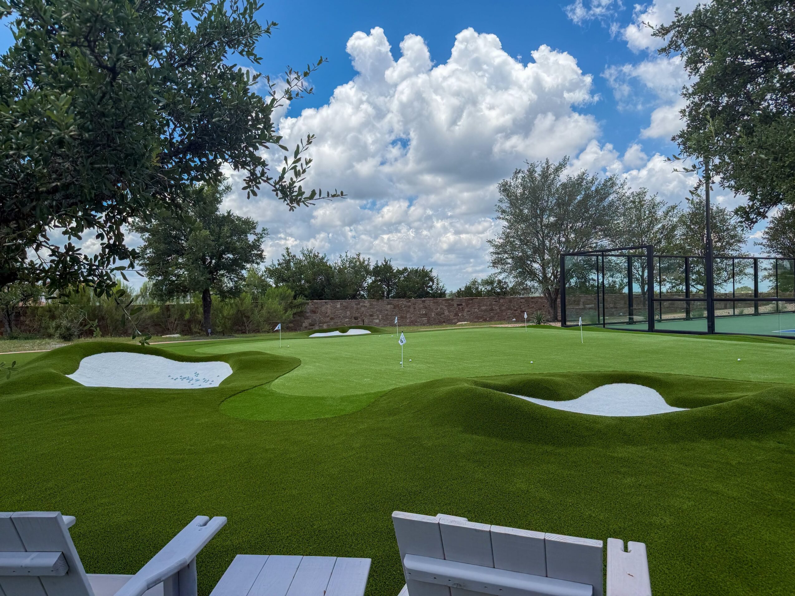 Green golf course with sand bunkers, trees, a fenced area, and patio chairs beneath a cloudy blue sky.