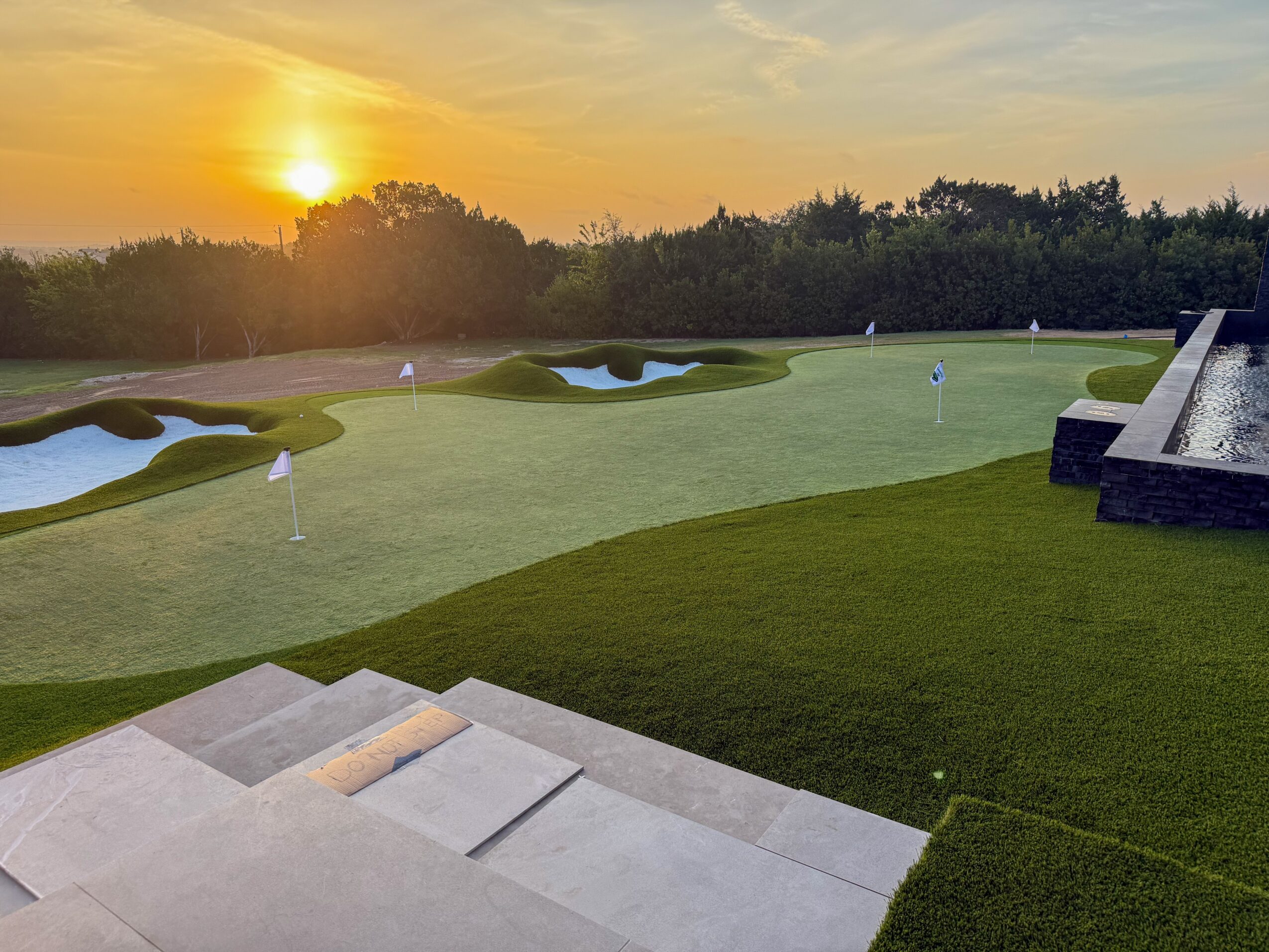 Sunset over a golf course with putting greens, sand bunkers, a water feature, and a stone terrace.