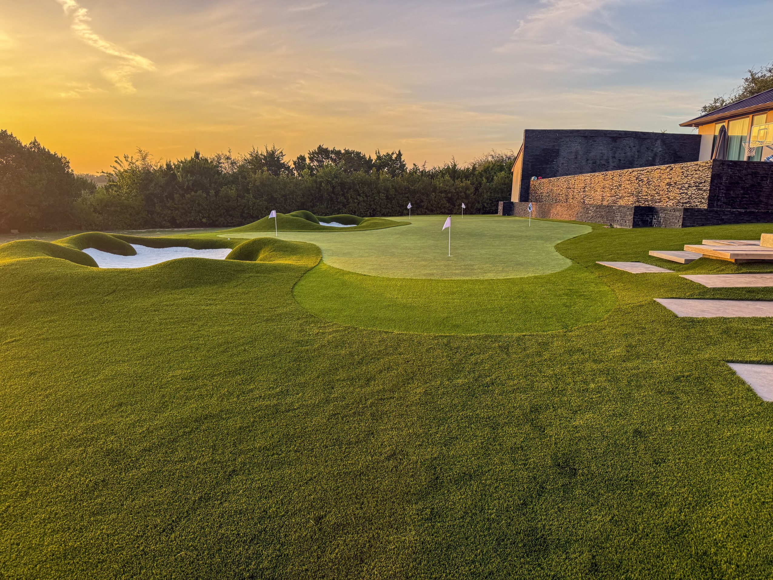 A manicured golf green with sand bunkers, a water feature, and a modern building at sunset.