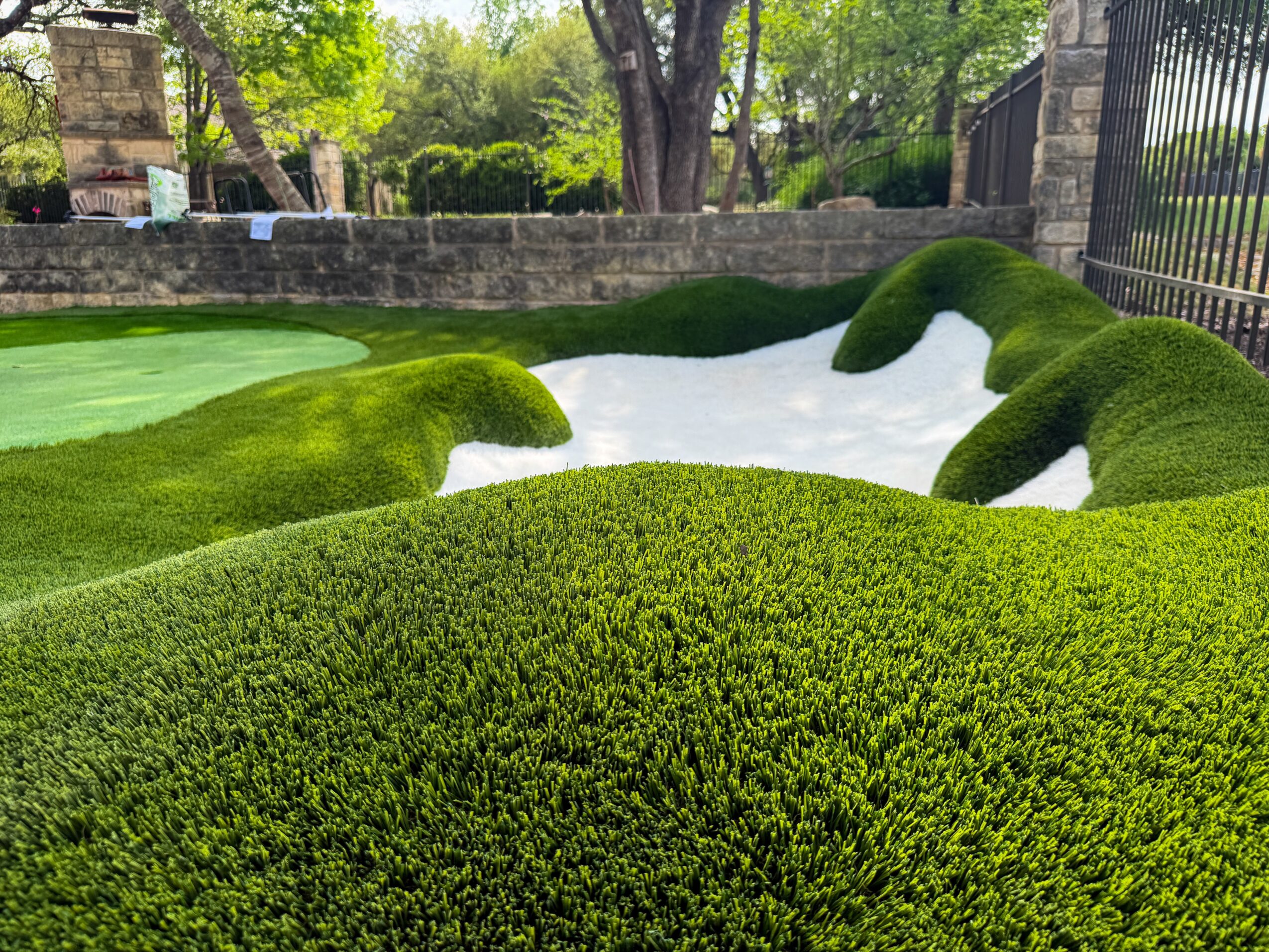 Sculpted green lawns and hedges surround a white gravel path in a garden bordered by a stone wall and iron fence.