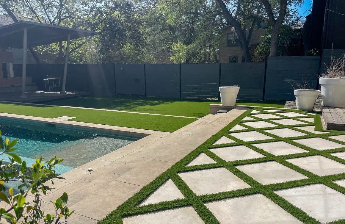 Outdoor pool beside a paved patio with geometric grass strips, potted plants, and trees.