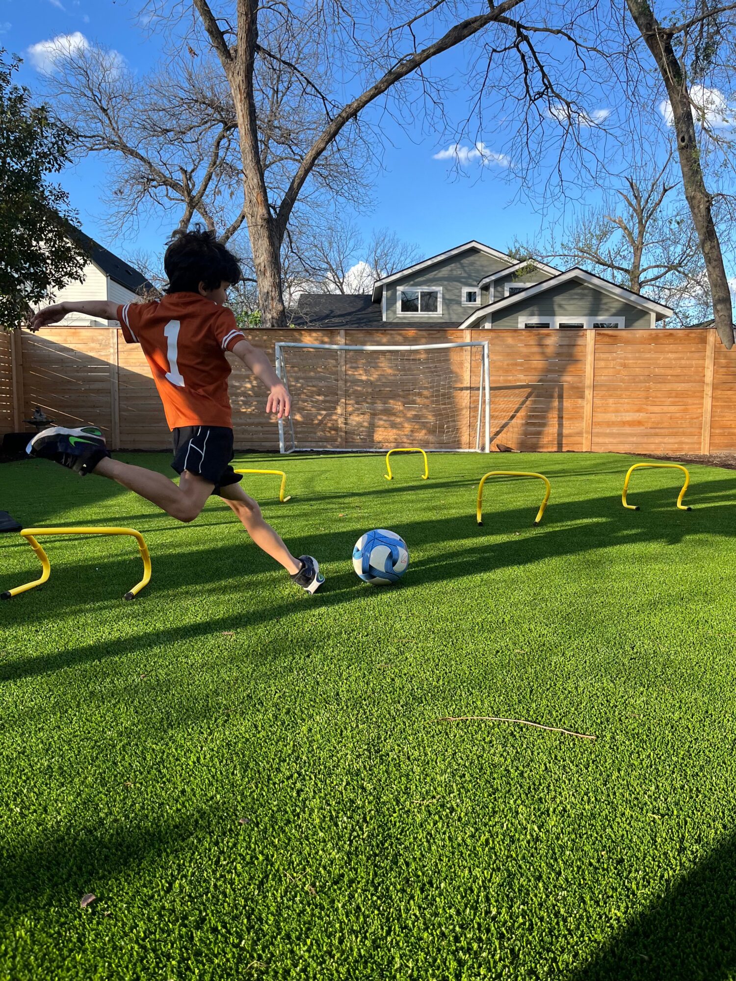 Person in an orange shirt runs toward a soccer ball on a grassy backyard field with training hurdles.