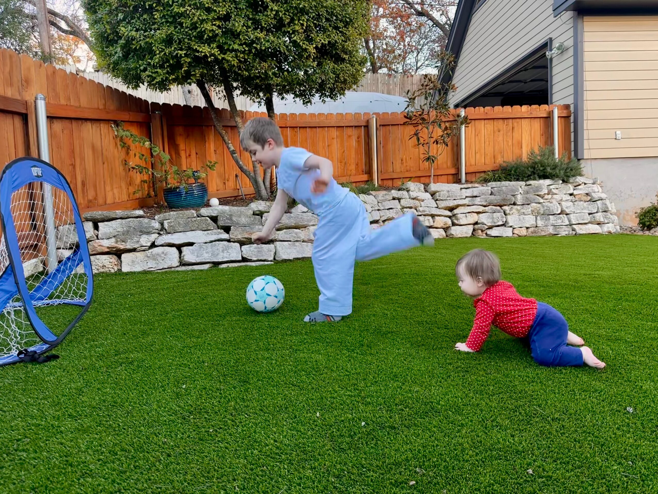 A child kicks a soccer ball in a backyard while a younger child crawls nearby.
