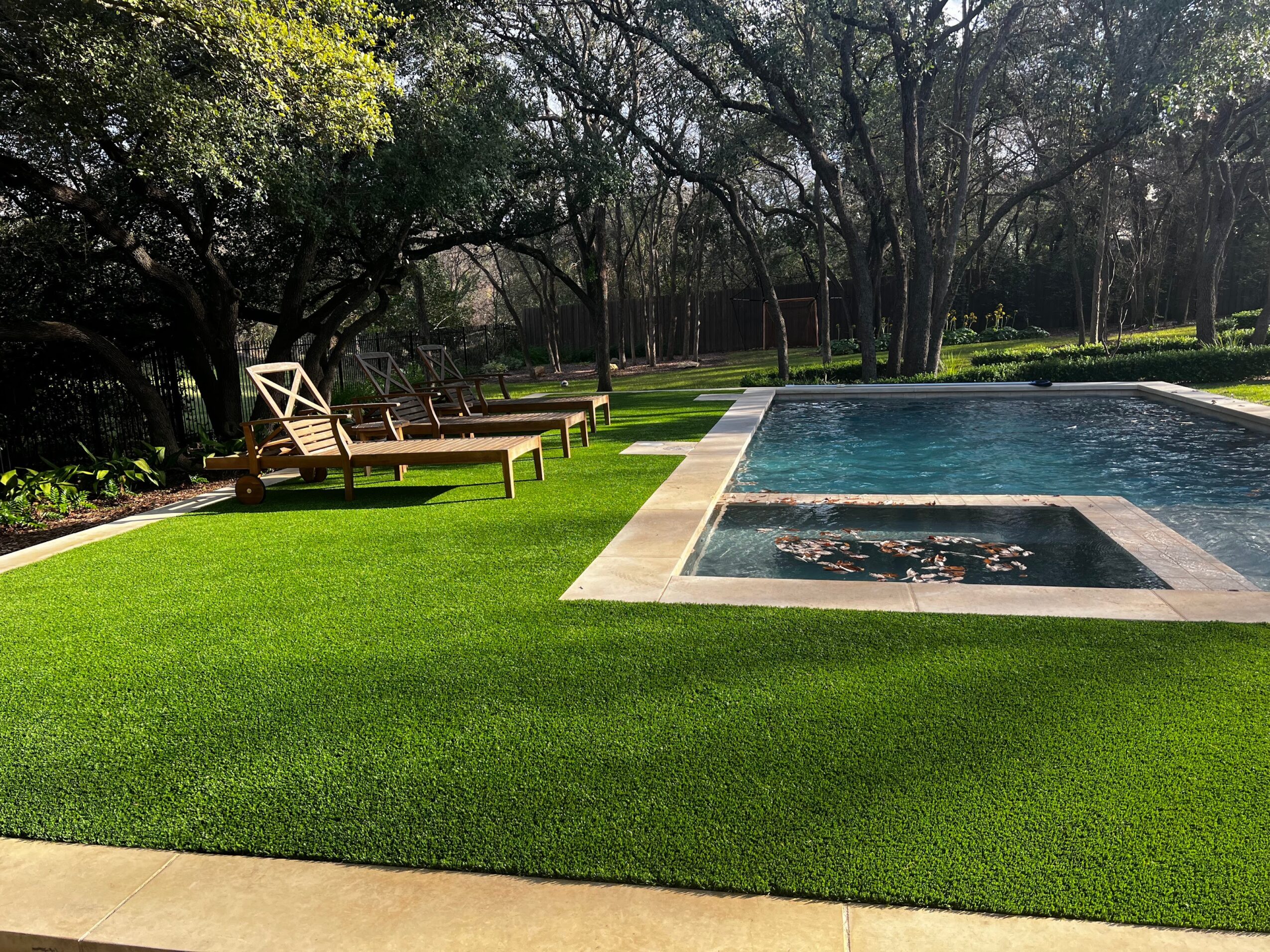 A lawn with wooden lounge chairs beside a rectangular swimming pool, surrounded by trees.