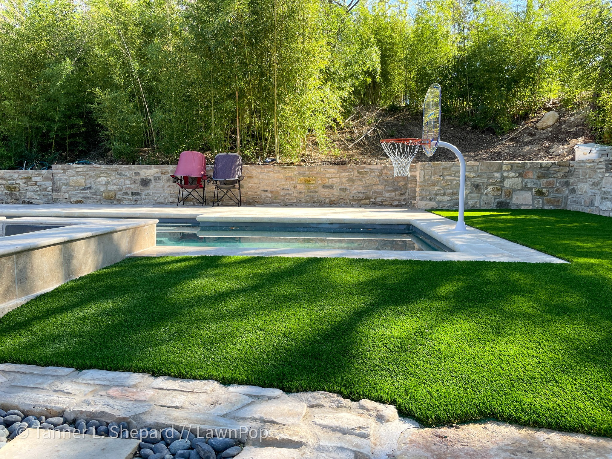 Outdoor pool beside a green lawn, with a basketball hoop, two chairs, stone walls, and trees.