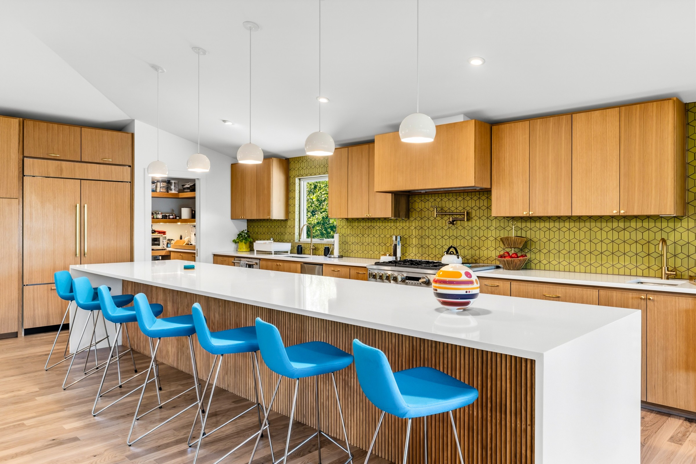 Modern kitchen with wooden cabinets, white island, blue chairs, green backsplash, and pendant lights.