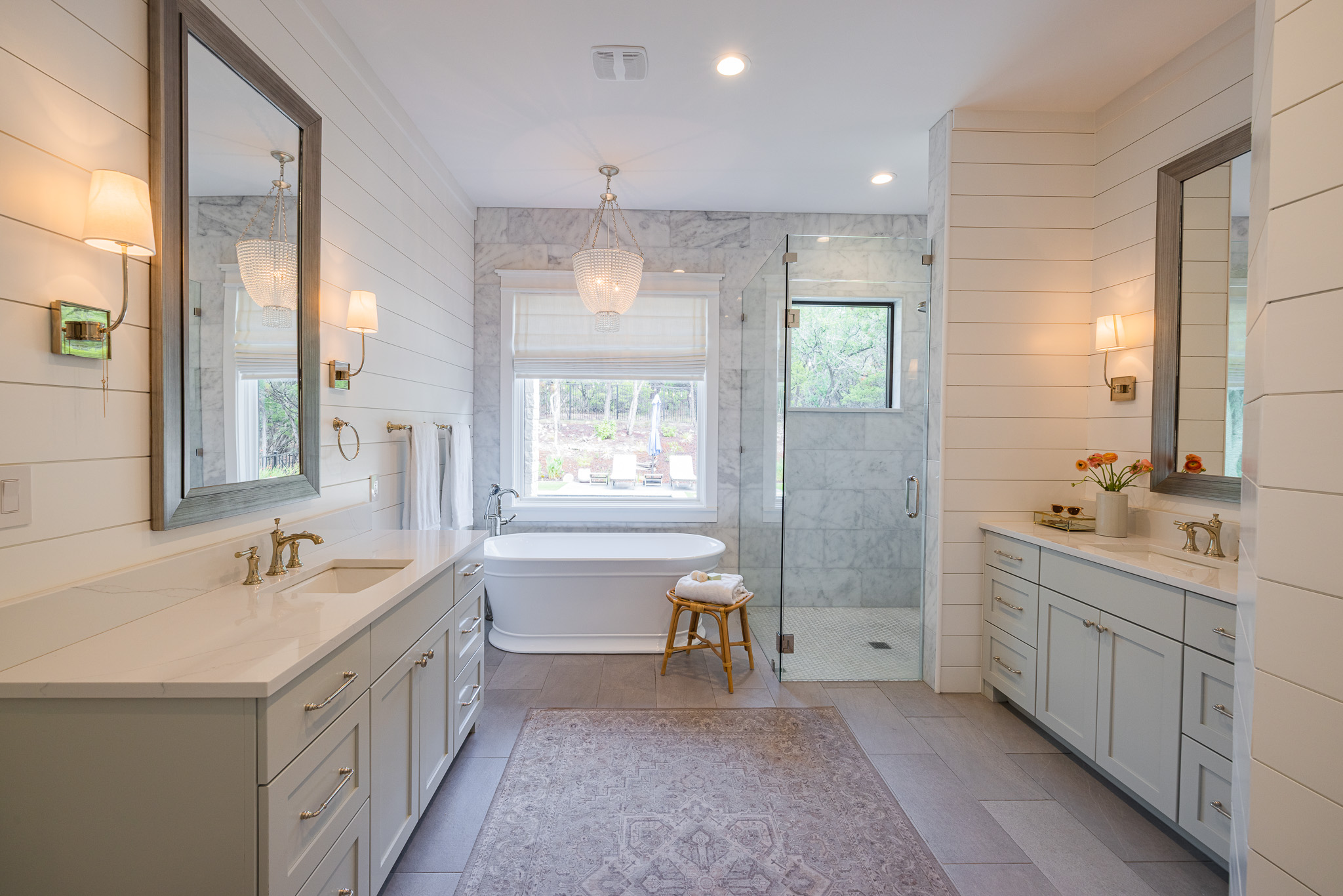Bathroom with double vanities, large mirror, bathtub, window, and a small stool, decorated in light colors.