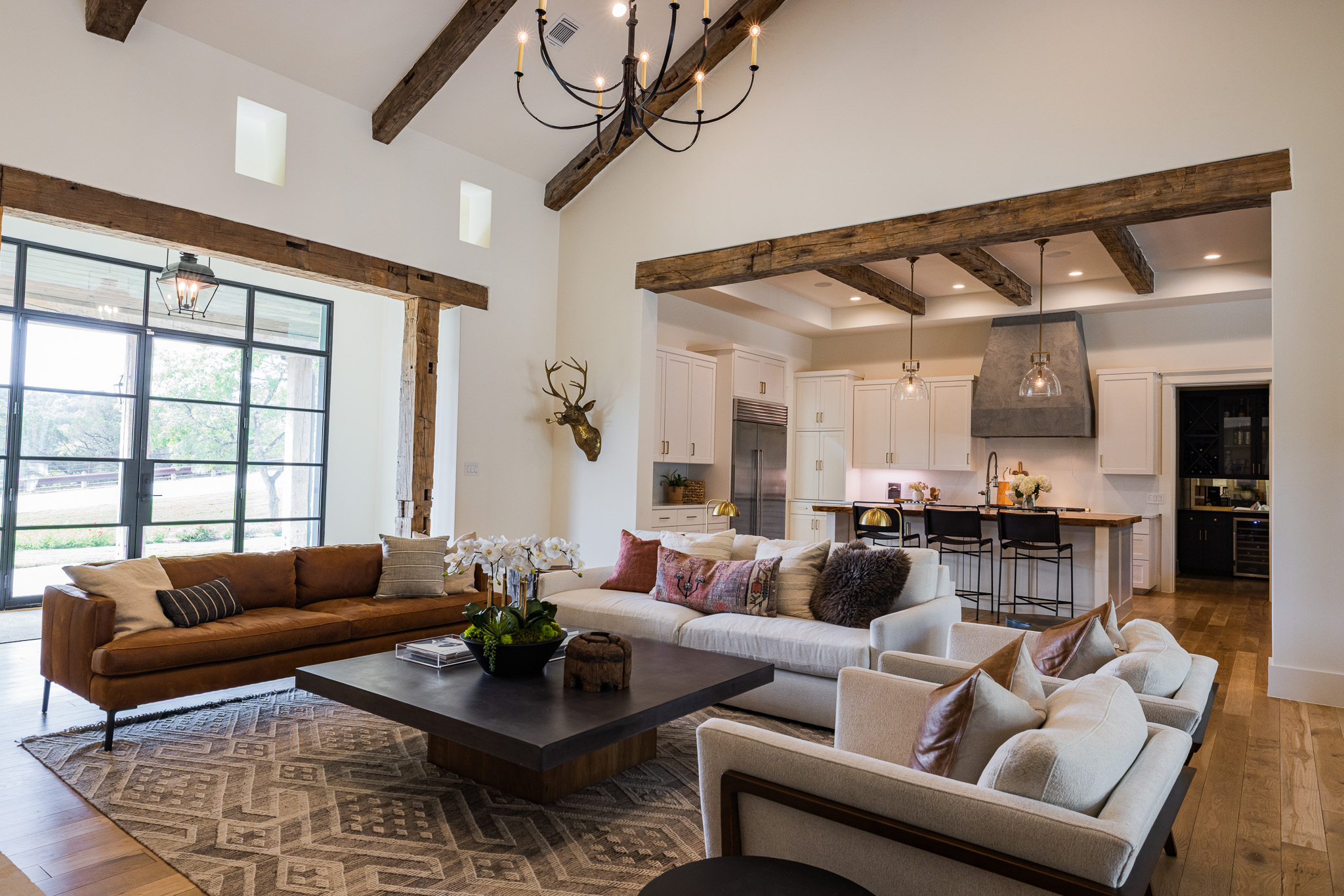 Living room with large window, brown and white sofas, wooden beams, and a chandelier, open to kitchen area.