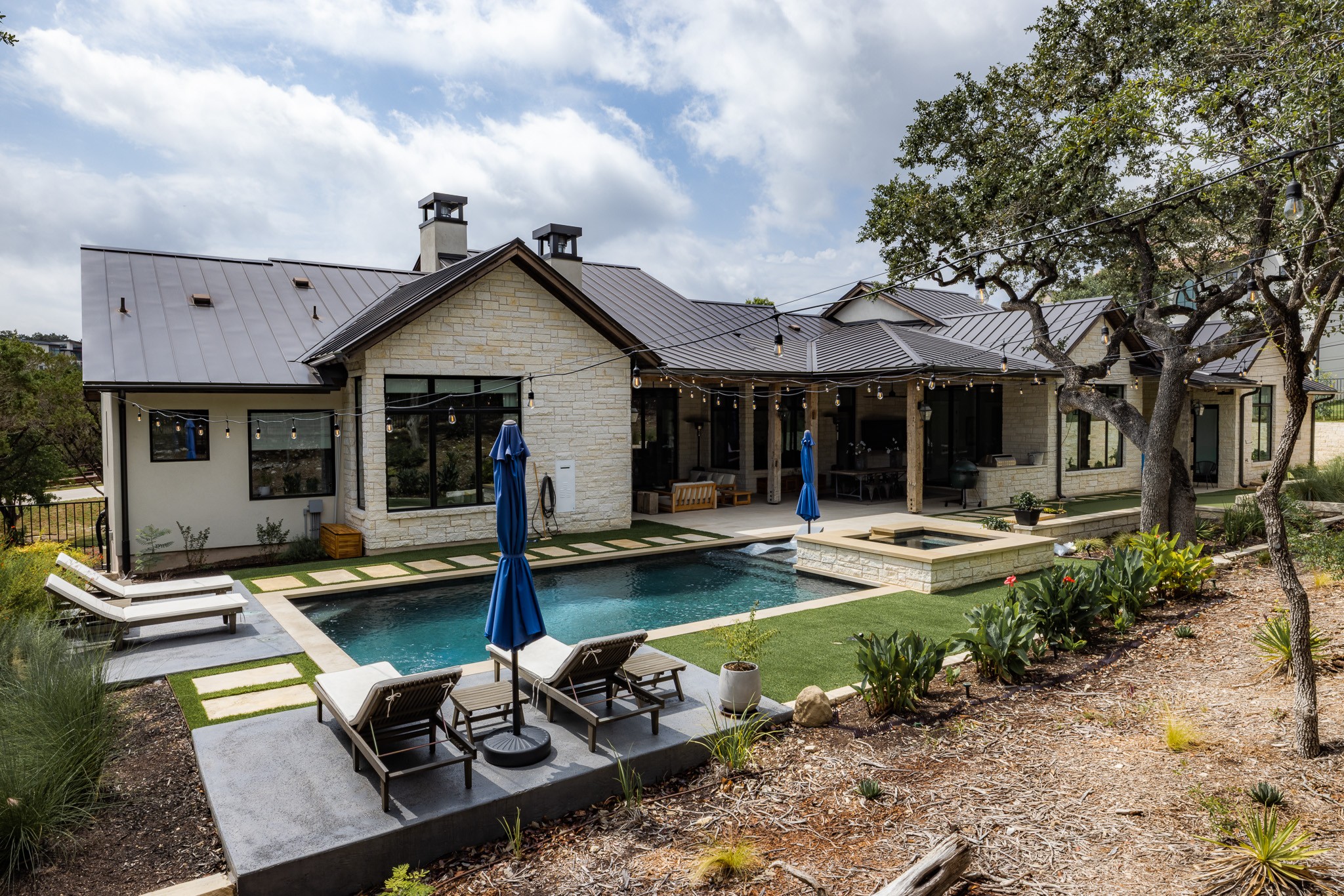 House with a backyard pool, outdoor seating, and trees, under a partly cloudy sky.