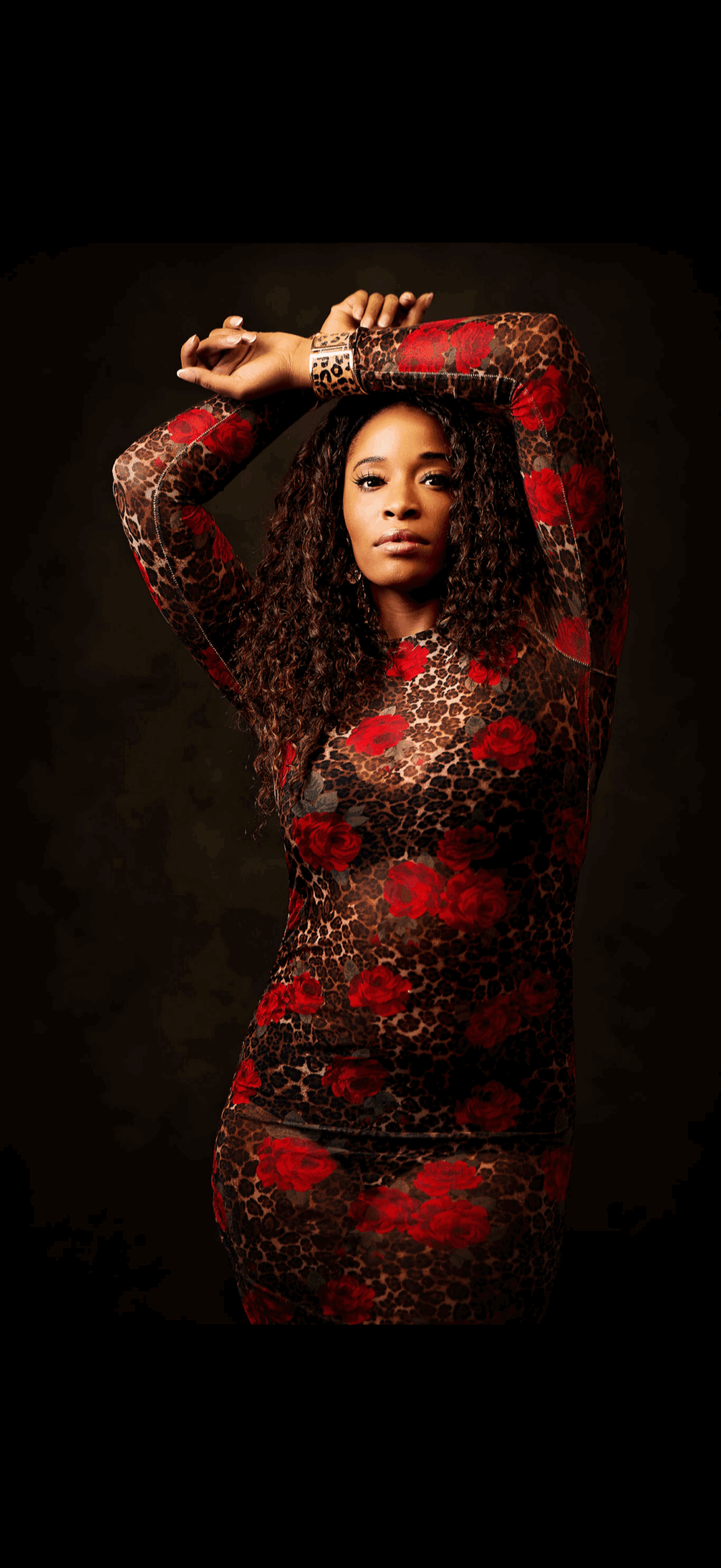 Woman with curly hair in a red and black patterned dress, arms raised above head, dark background.
