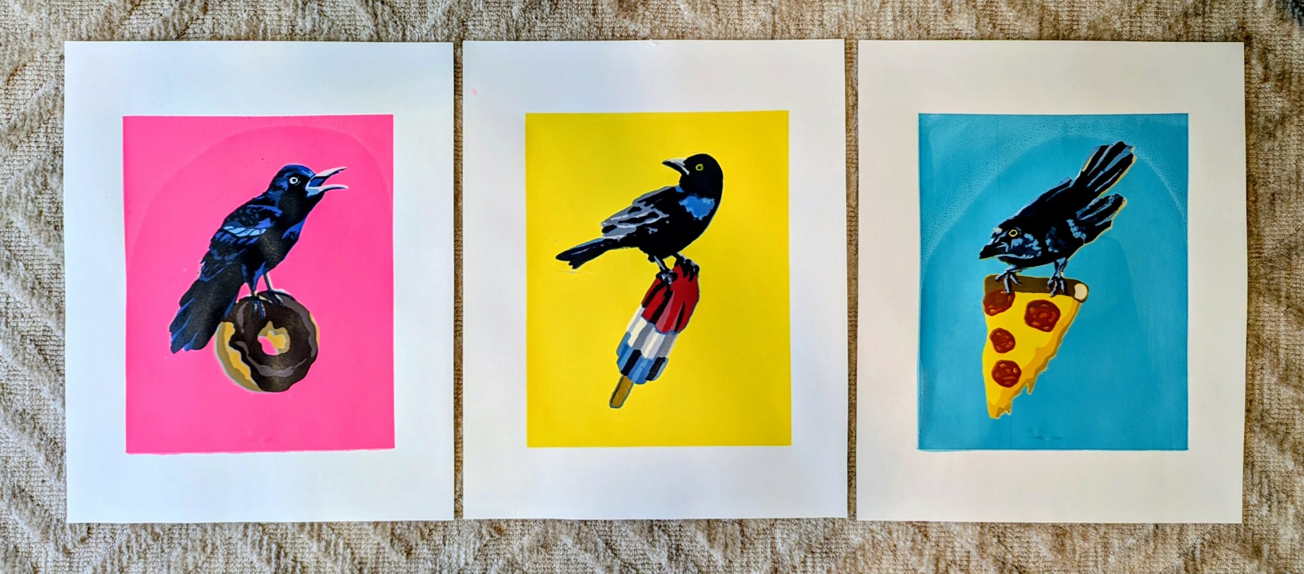 Three bird illustrations, each holding a different food item, on colorful backgrounds, displayed on a carpeted surface.