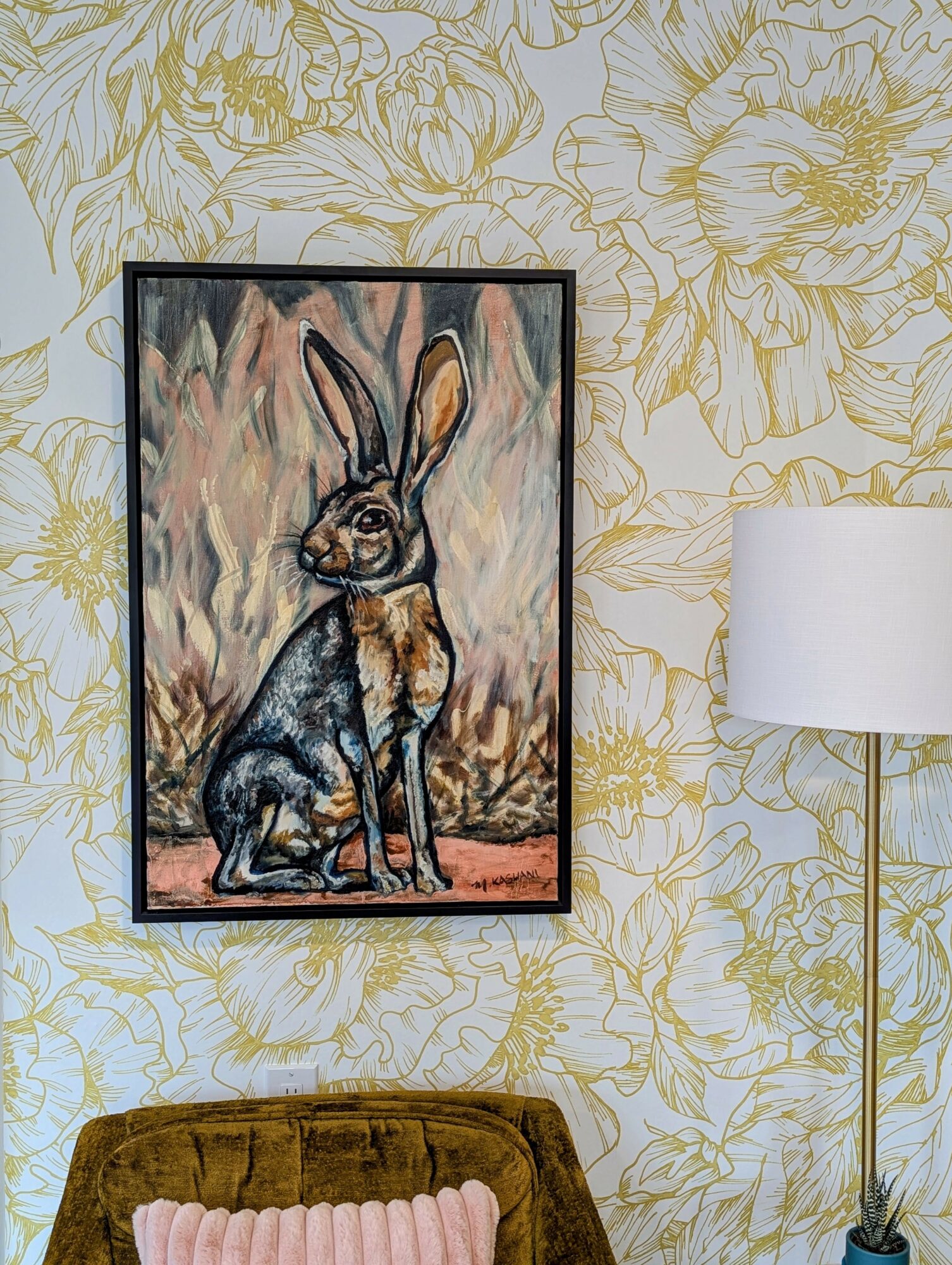 Painting of a hare with tall ears, sitting outdoors, framed on a floral wallpaper background, next to a white lamp.