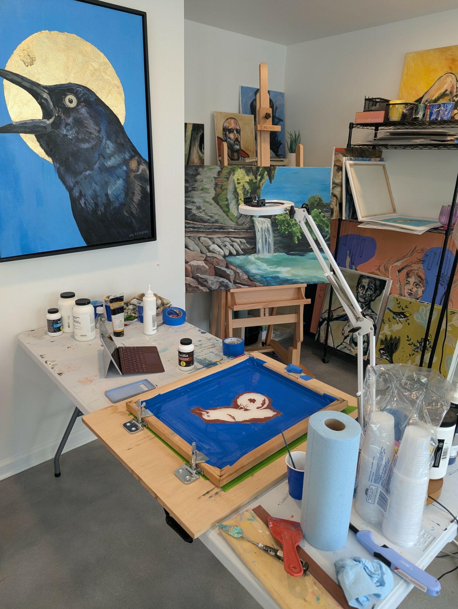 Art studio with paintings, supplies, and a work-in-progress piece on a table, including paint bottles, paper towels, and tools.