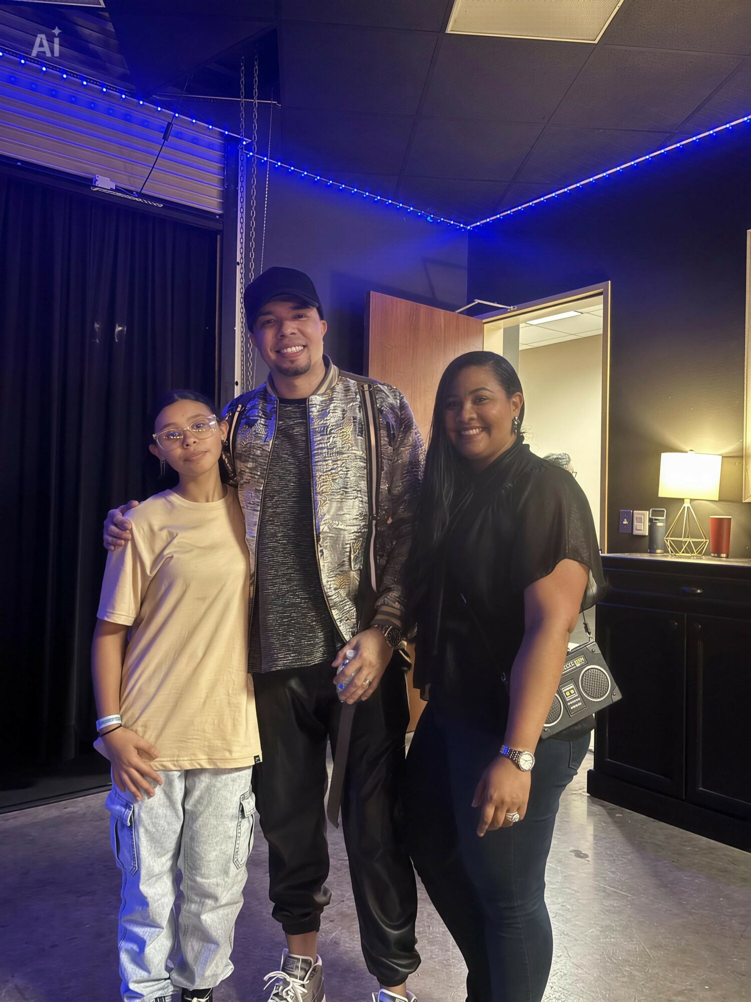 Three people standing together indoors, smiling, with blue LED lights and dark curtains in the background.