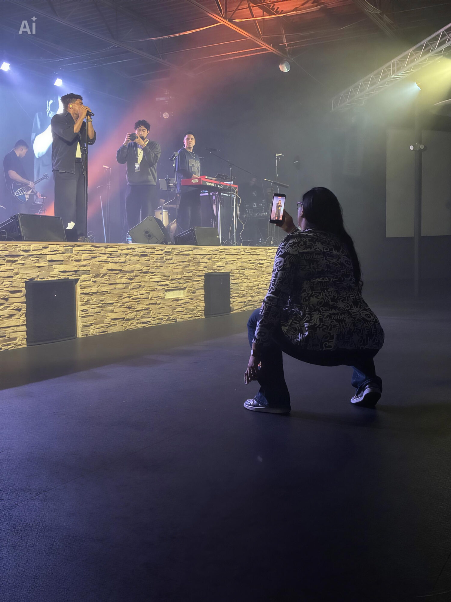 Person crouching in front of stage with performers, stage lights, and equipment visible, audience area in foreground.