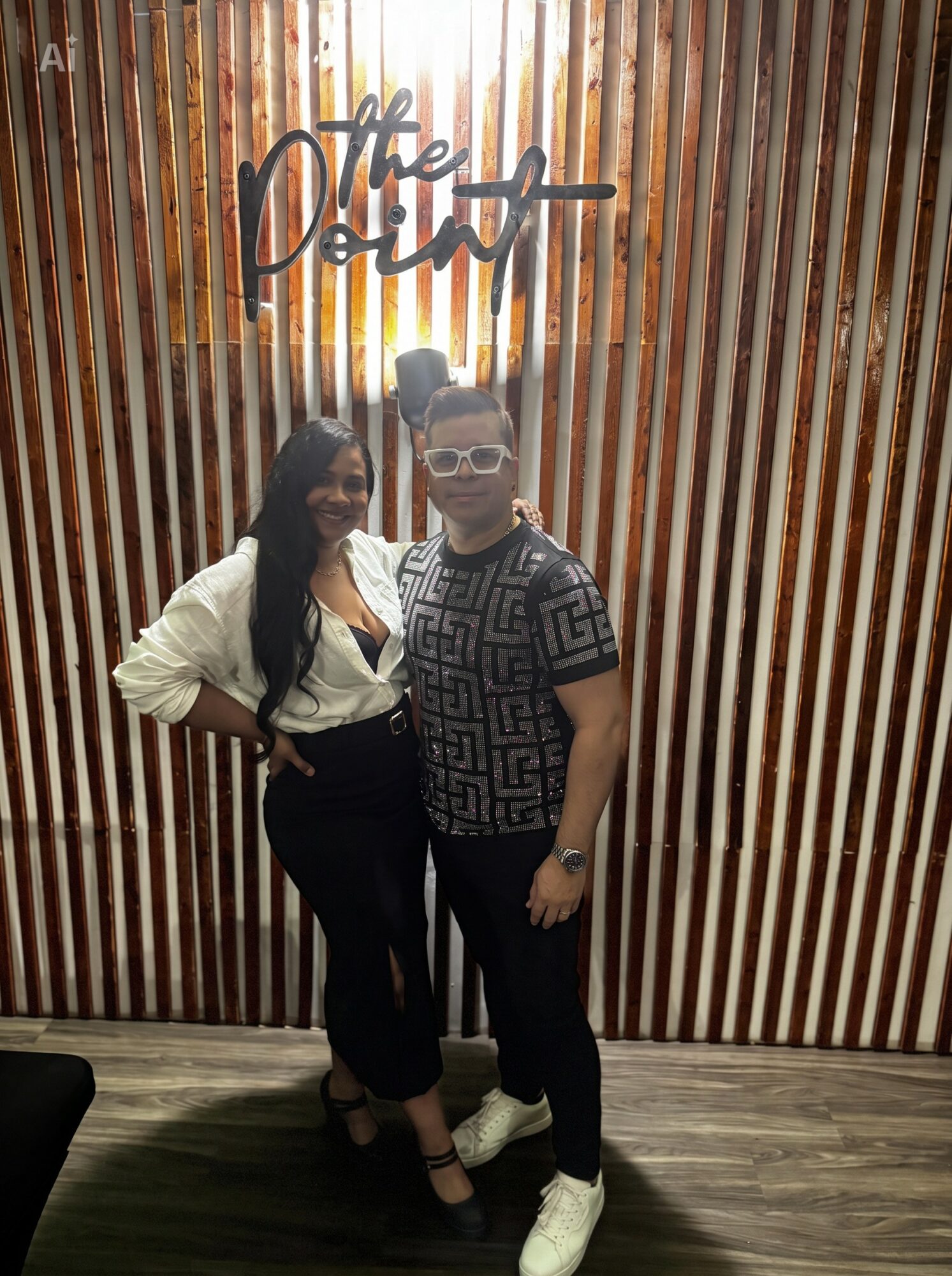Two women stand in front of a wooden wall with a neon sign that says 'The Dine'. One woman wears a white blouse and black pants, the other wears a patterned shirt and glasses.