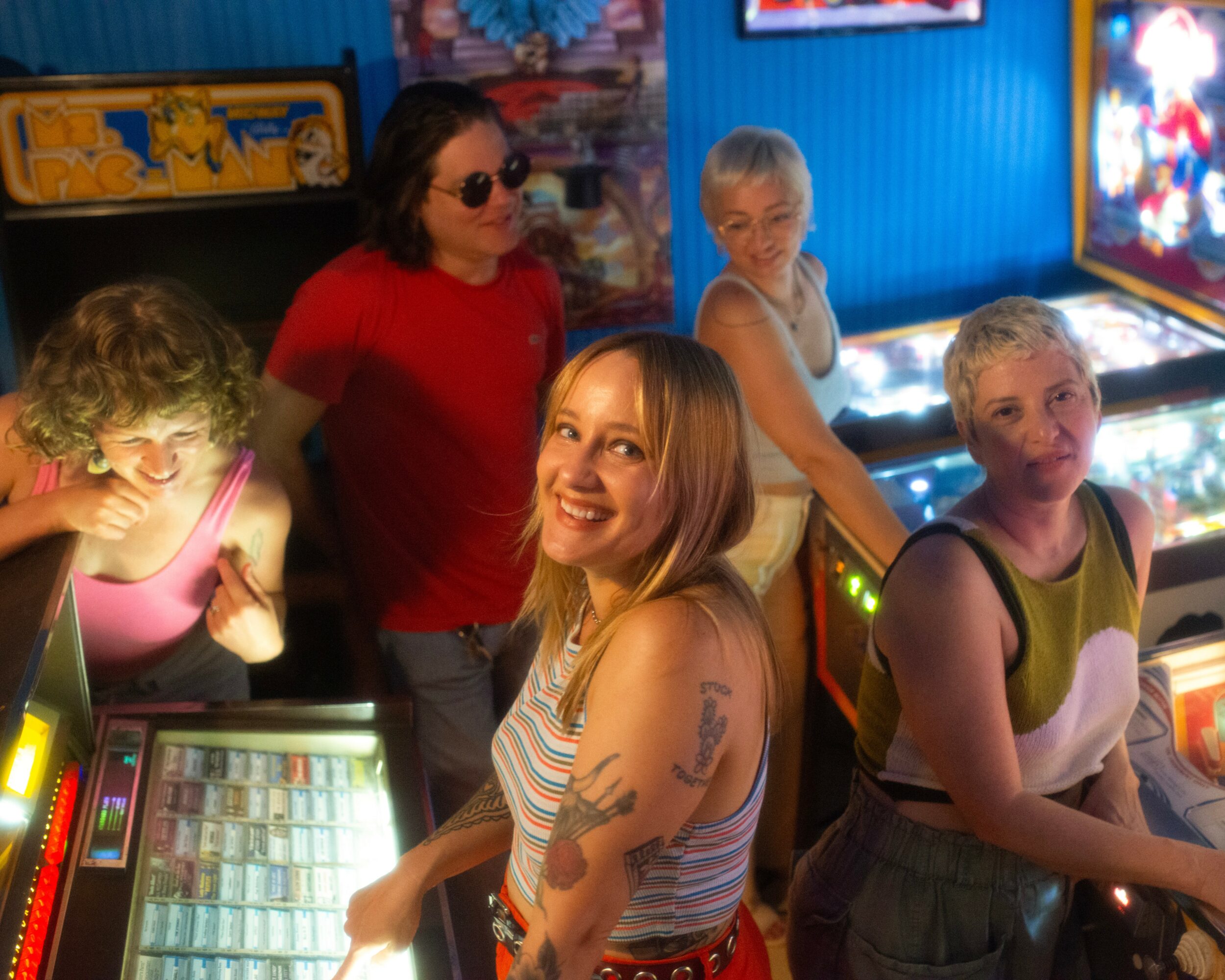 Five women are in an arcade, some playing games and smiling, with colorful screens and decorations around them.